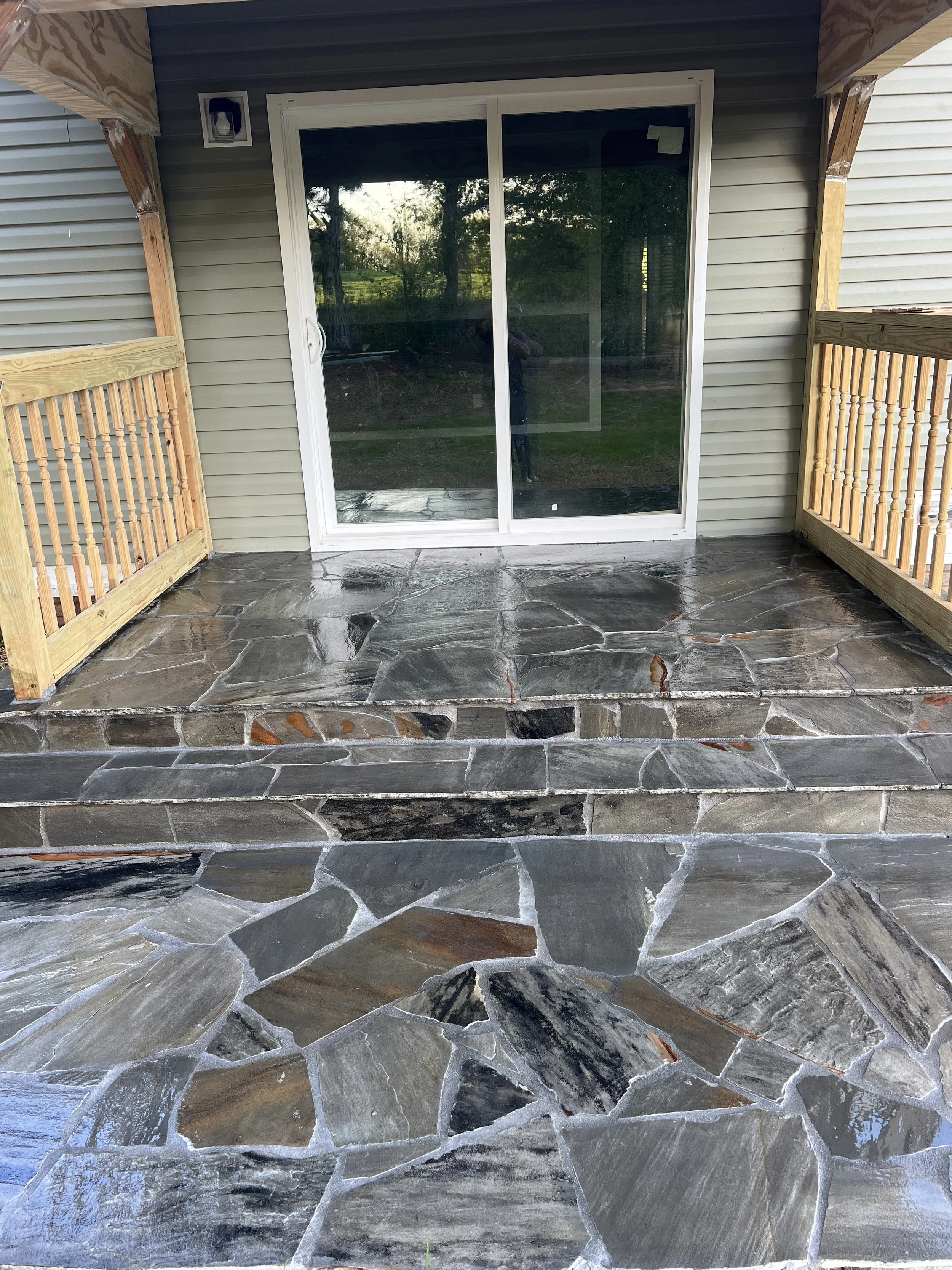 A small porch with stone tile flooring and sliding glass door leading into a house. The porch is enclosed by wooden railings on both sides.