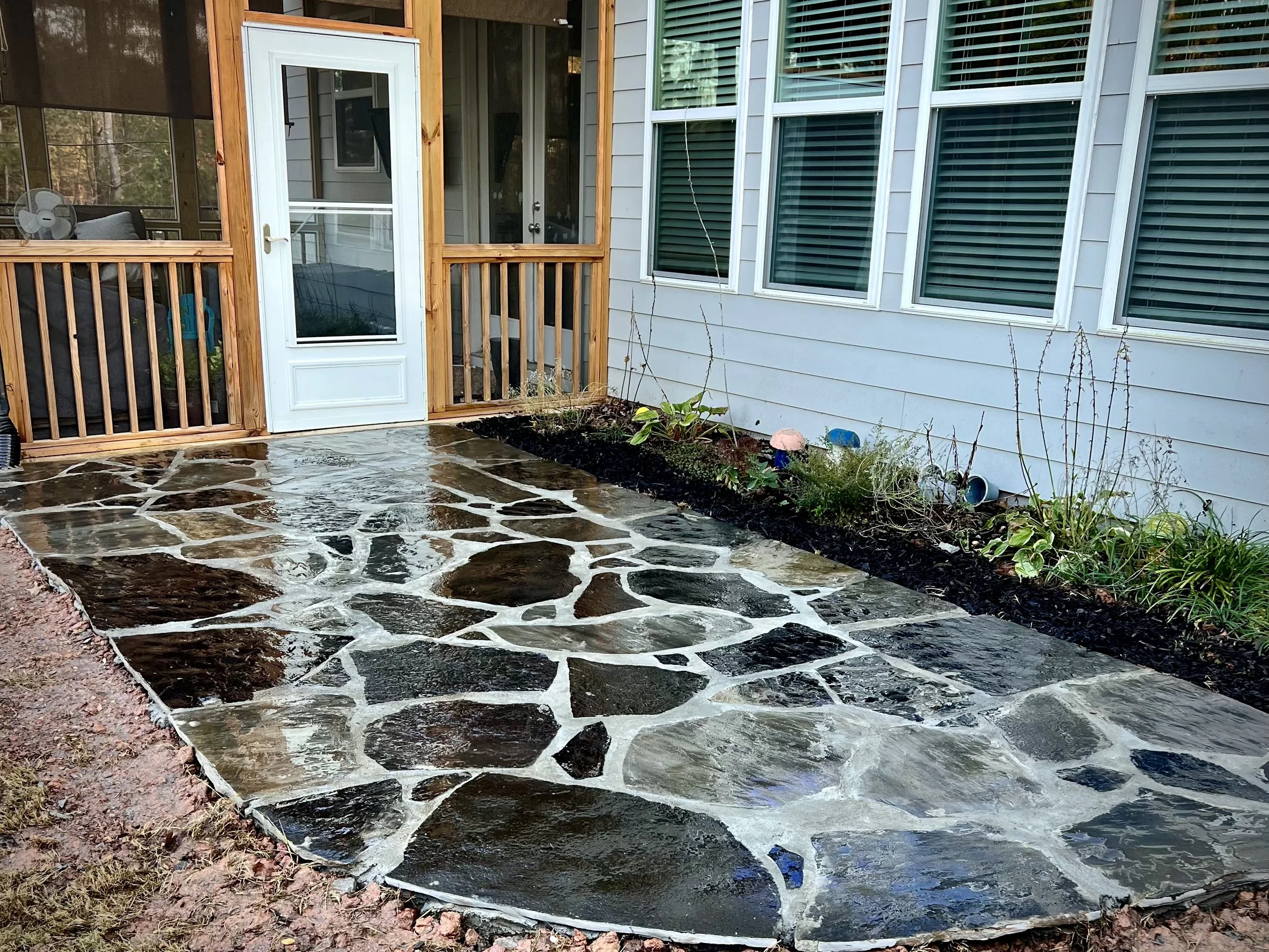 Stone patio outside a house with a white door, a screened porch, and a flower bed with plants along the house.