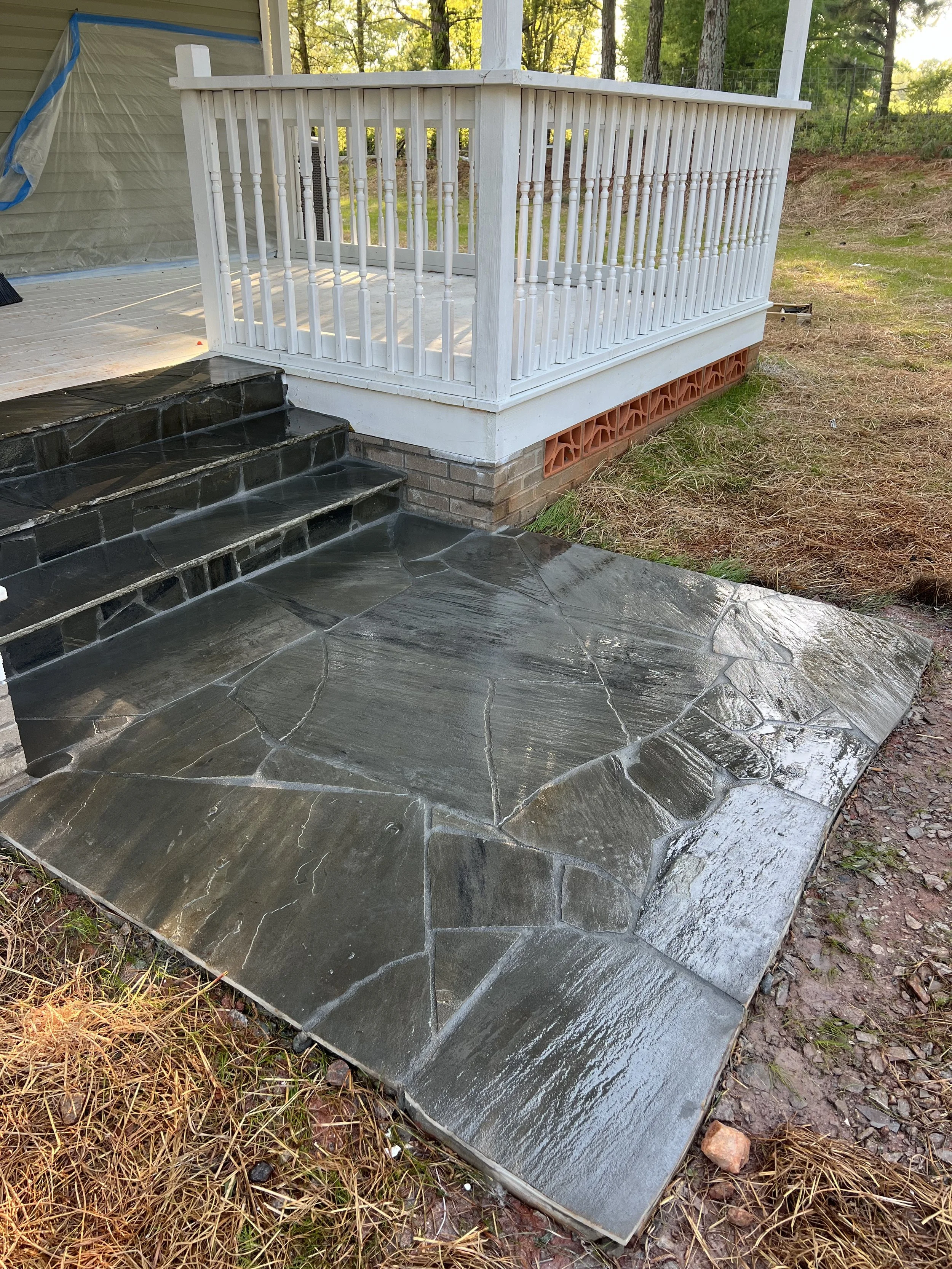 Black stone patio with steps leading to a white wooden porch with railing, outdoor setting with trees and grass.