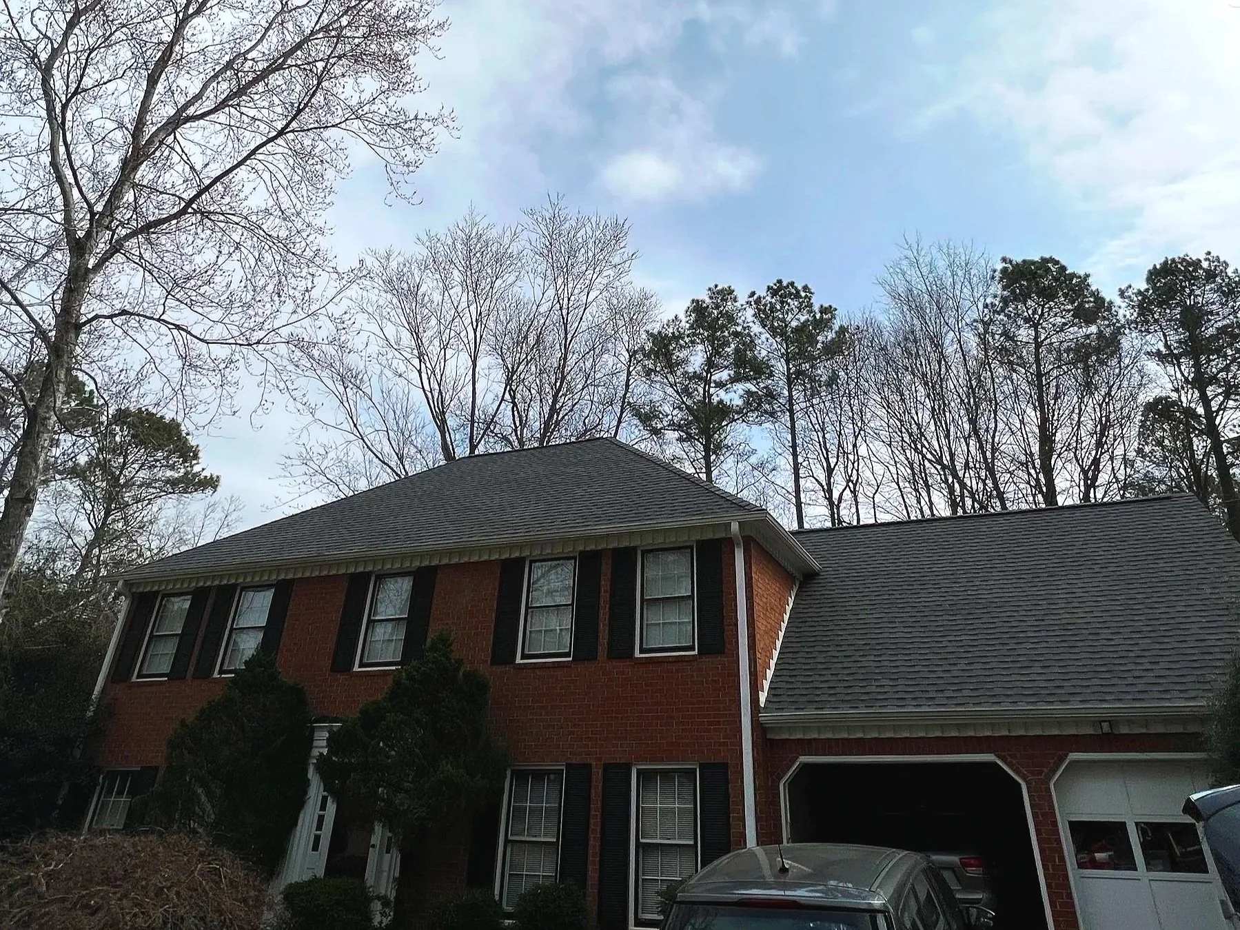 Two-story brick house with black shutters, green bushes in front, white garage doors, parked cars, leafless trees in the background, cloudy sky.