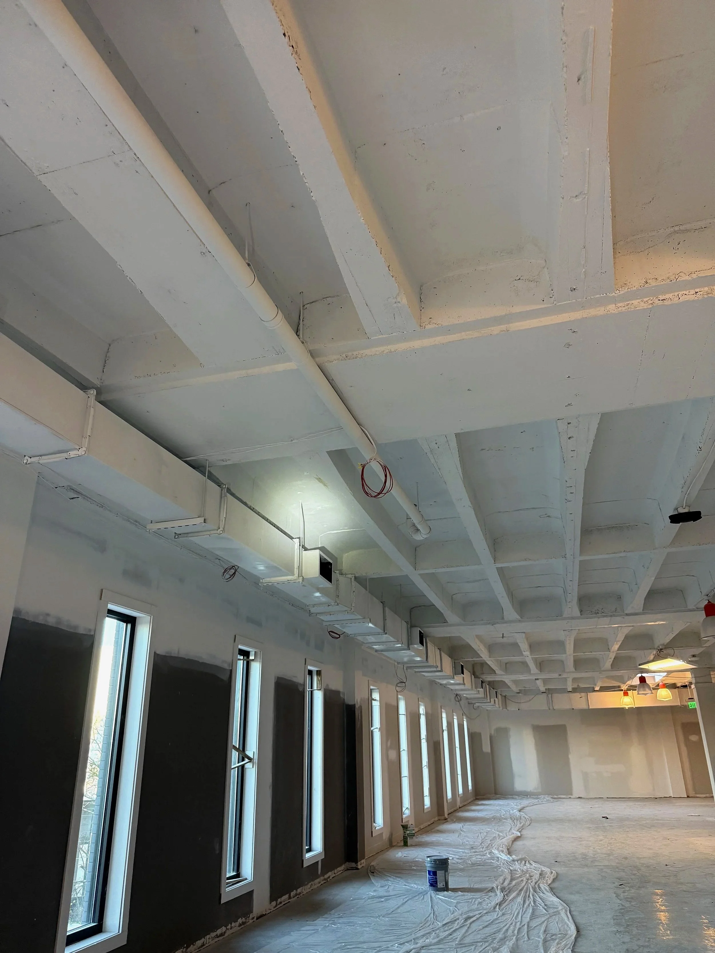 Interior of a building under construction with partially finished walls, exposed ceiling with beams and pipes, and tall narrow windows with drapes or coverings.