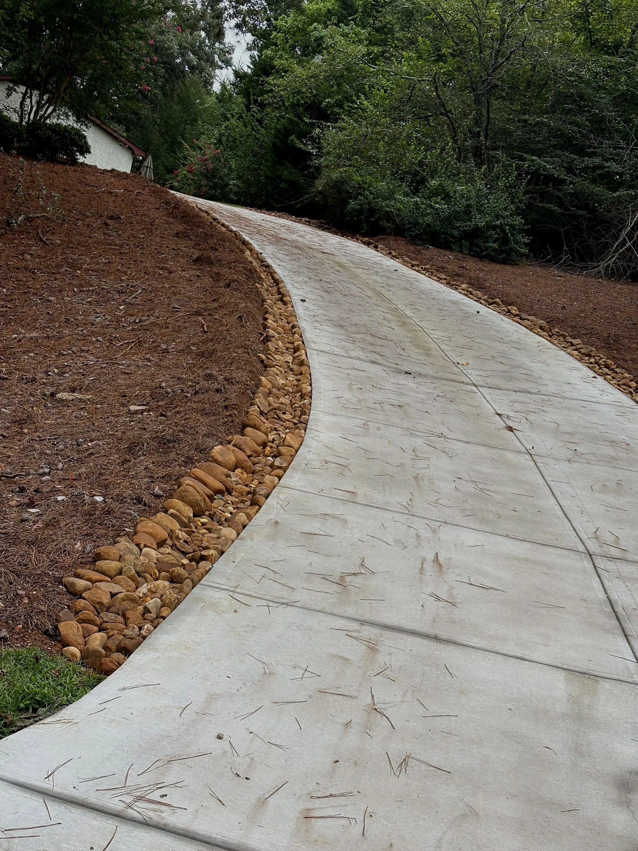 Curving concrete pathway lined with small rocks, on a slope with dirt on one side and trees on the other.