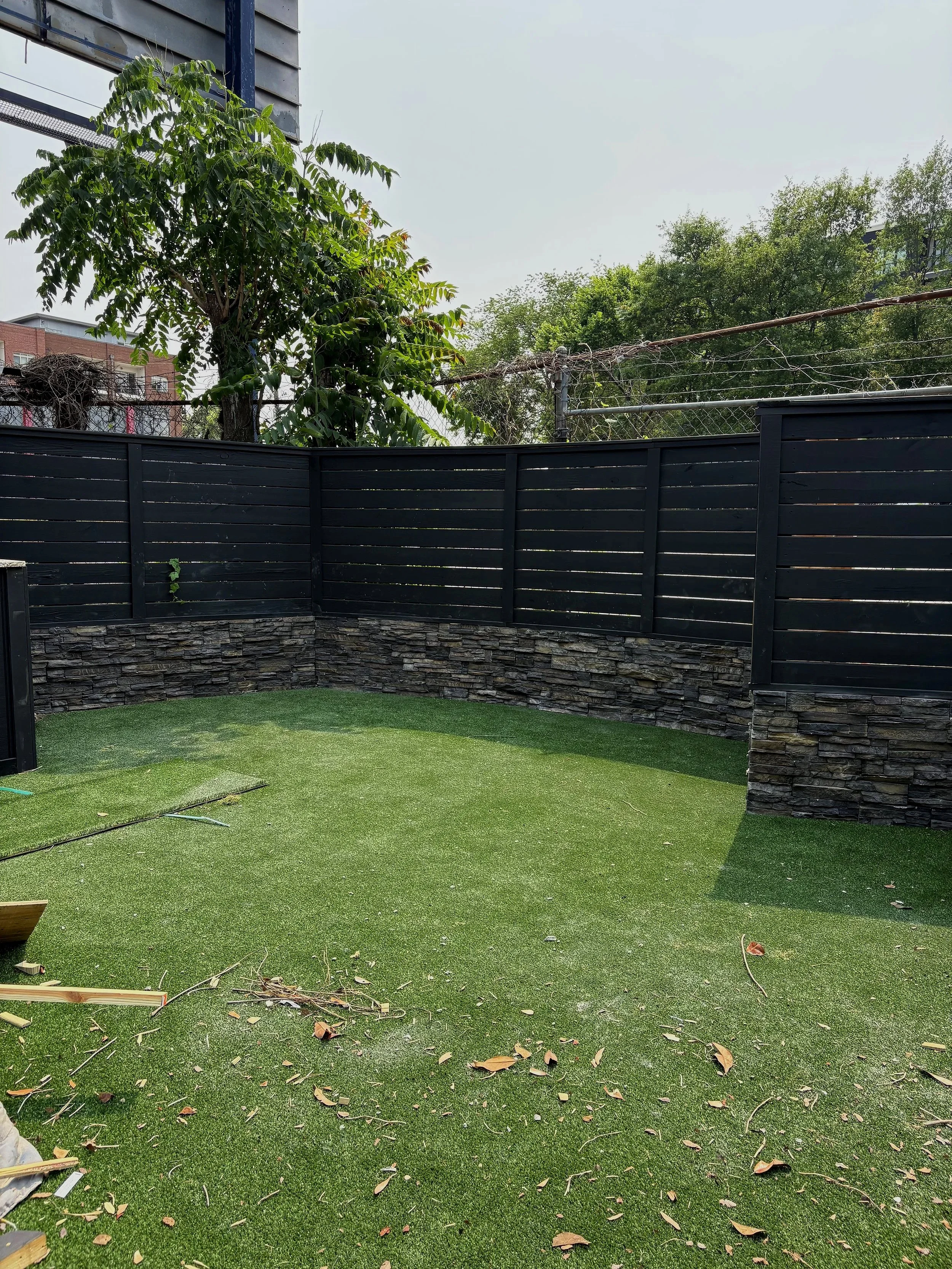 A backyard with artificial grass, a black wooden fence with stone veneer at the bottom, and a few scattered leaves and small debris on the ground.