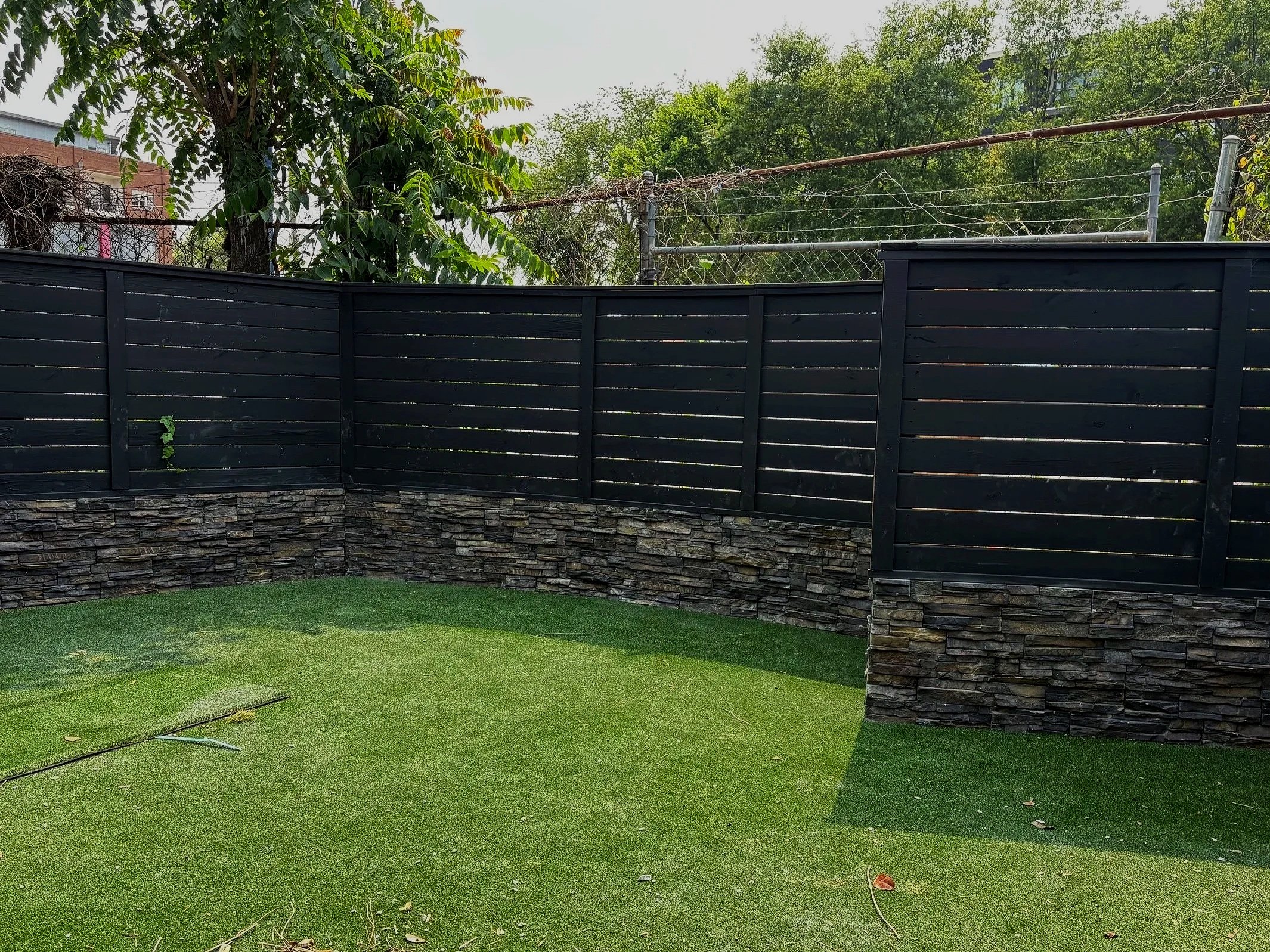 A backyard with green artificial grass, black wood fence with stone base, and trees in the background.