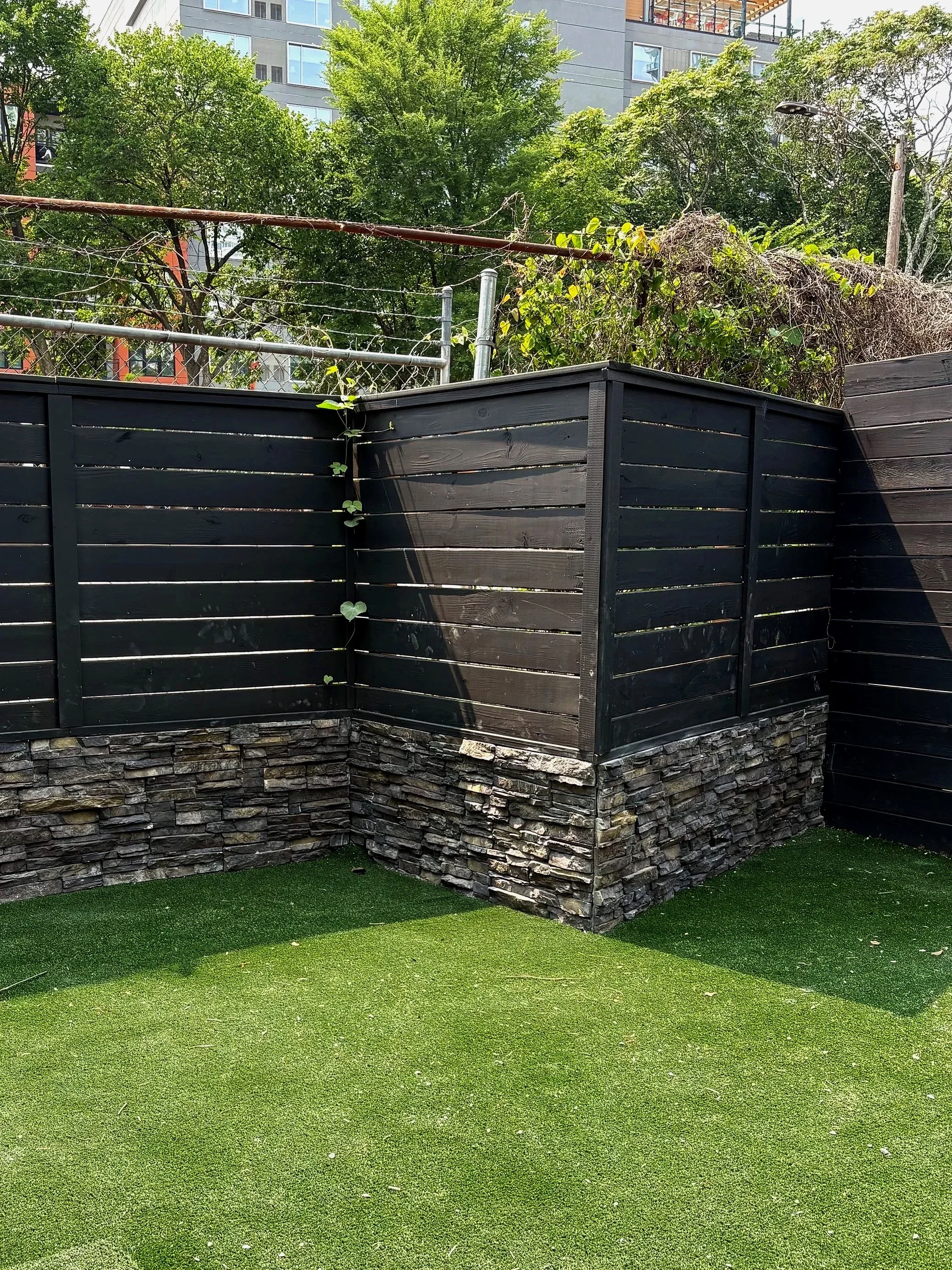 Empty outdoor area with artificial grass, a black wooden fence with a stone base, and trees in the background. A building with the word 'REVER' partially visible is in the distance.