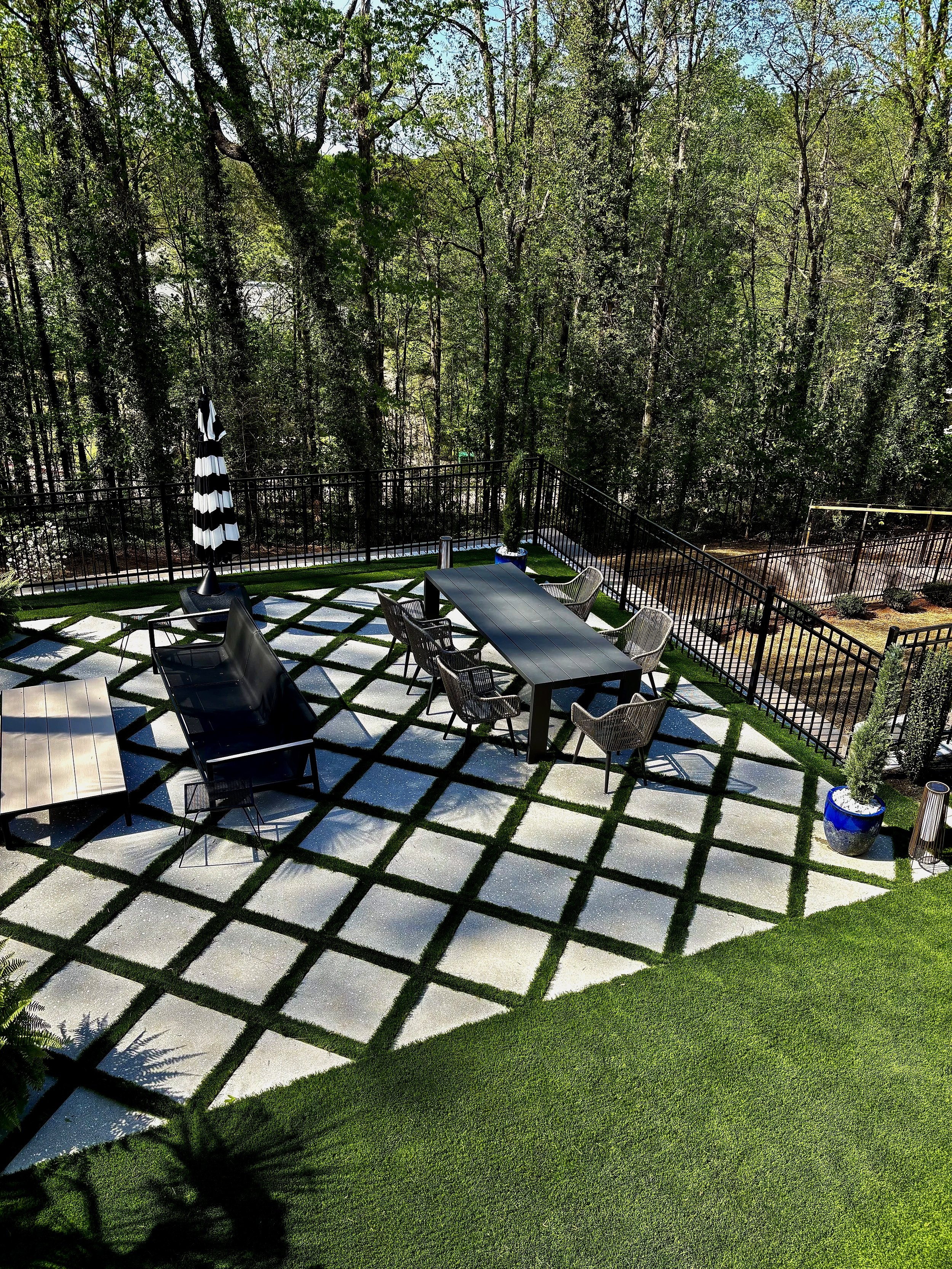 An outdoor patio area with a modern black dining table and six chairs, a large black umbrella, a black lounge chair, and decorative potted plants, surrounded by a black metal fence and lush green trees.