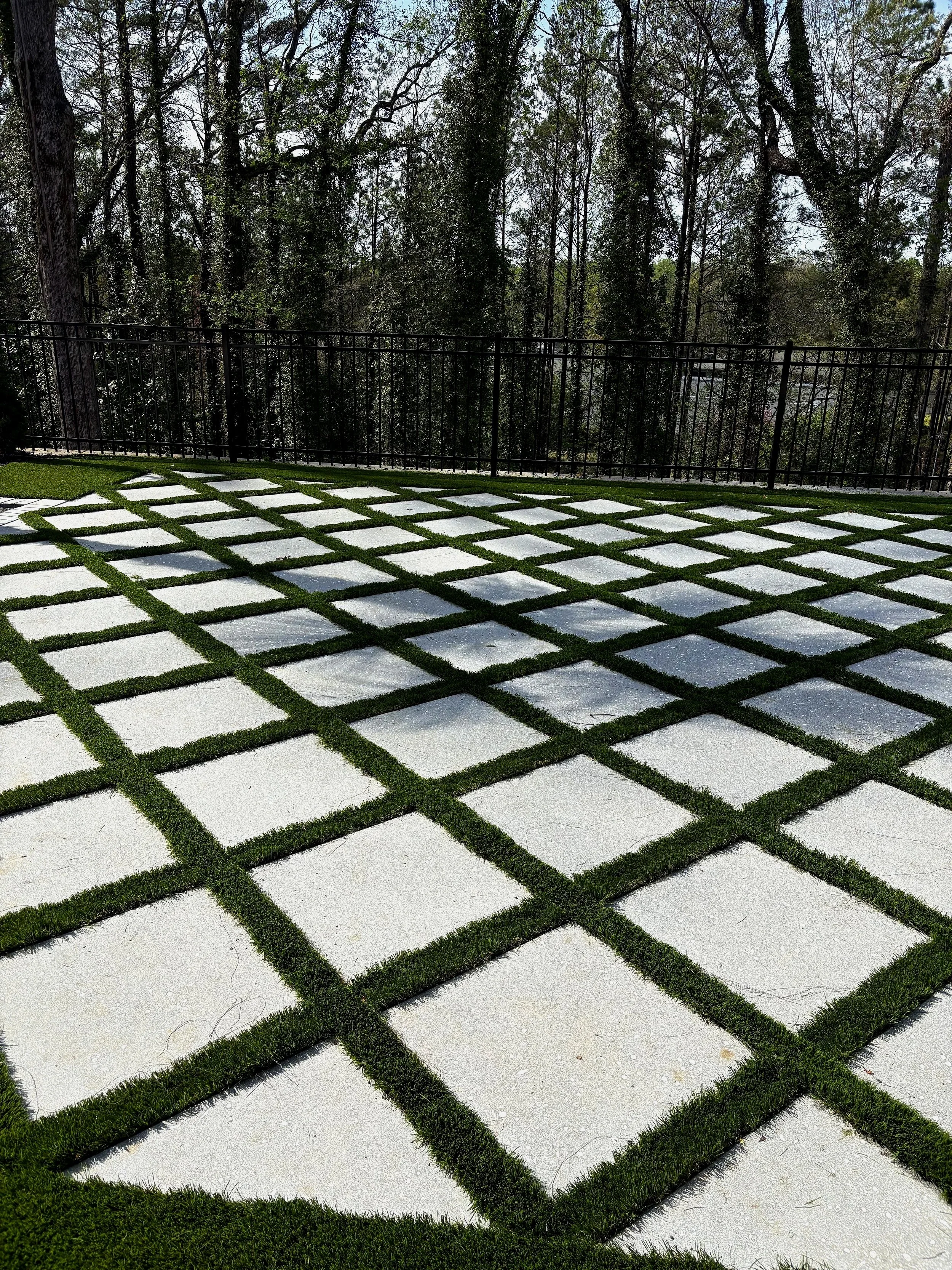 Outdoor patio with white paving squares separated by green grass, surrounded by a black metal fence and tall trees in the background.