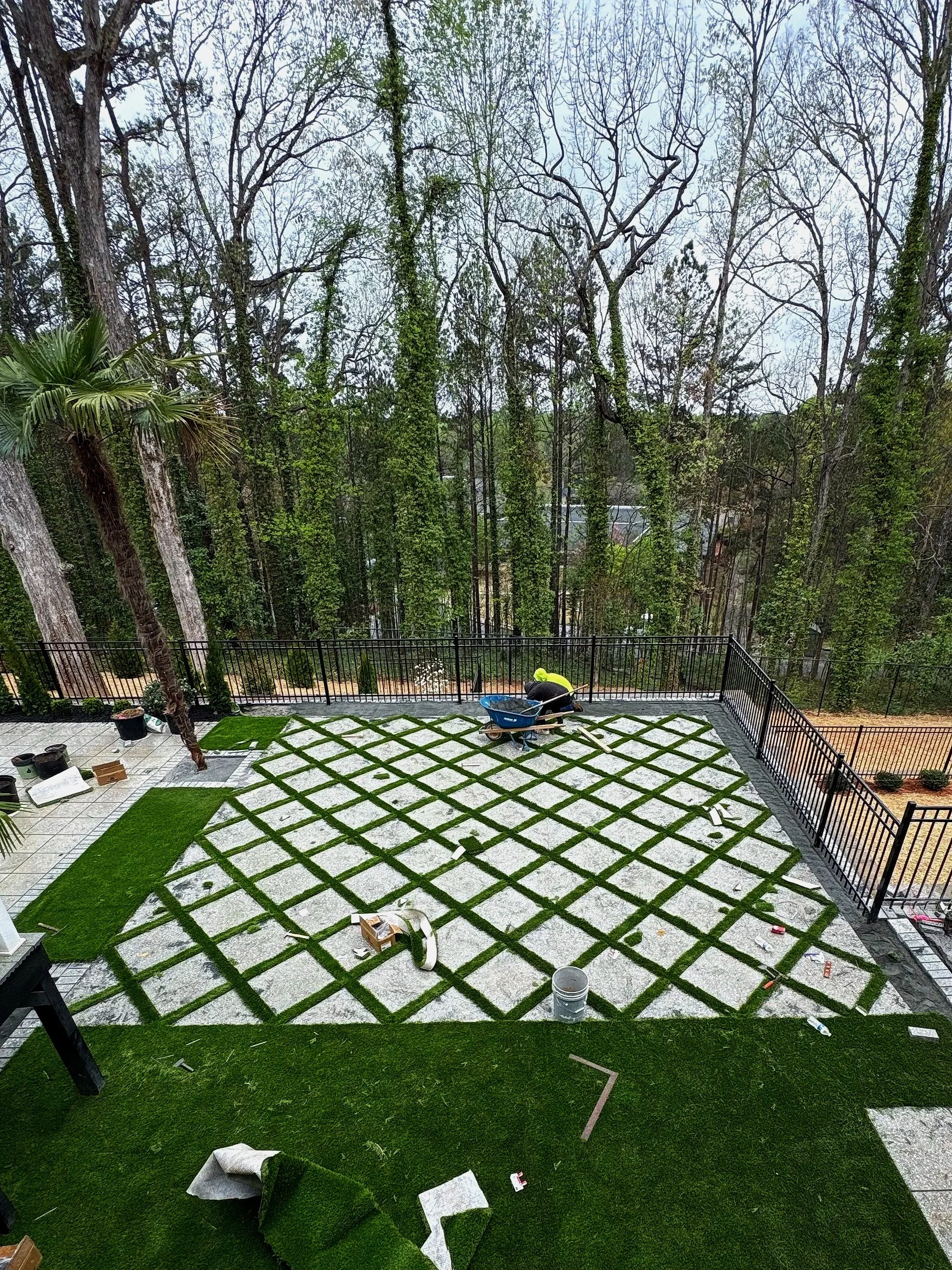 A worker installing a decorative grid pattern of grass and concrete on an outdoor patio area, with trees in the background, and tools and supplies scattered around.