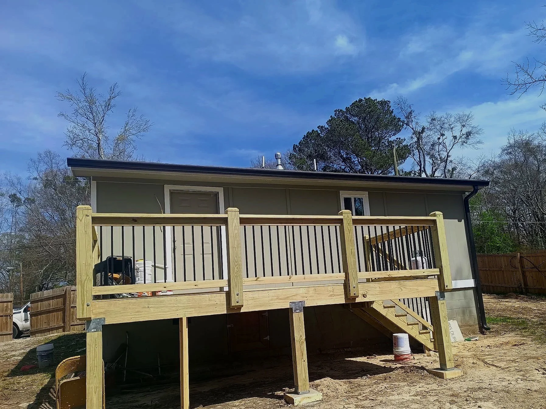 New wooden deck being built on the back of a house with outdoor stairs and a railing, on a dirt backyard with a fence and trees.