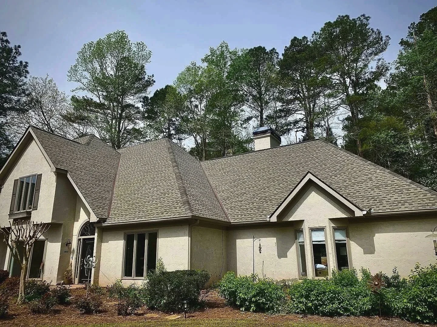 A large house with a beige exterior and gray shingled roof, surrounded by trees and bushes, with a clear sky overhead.