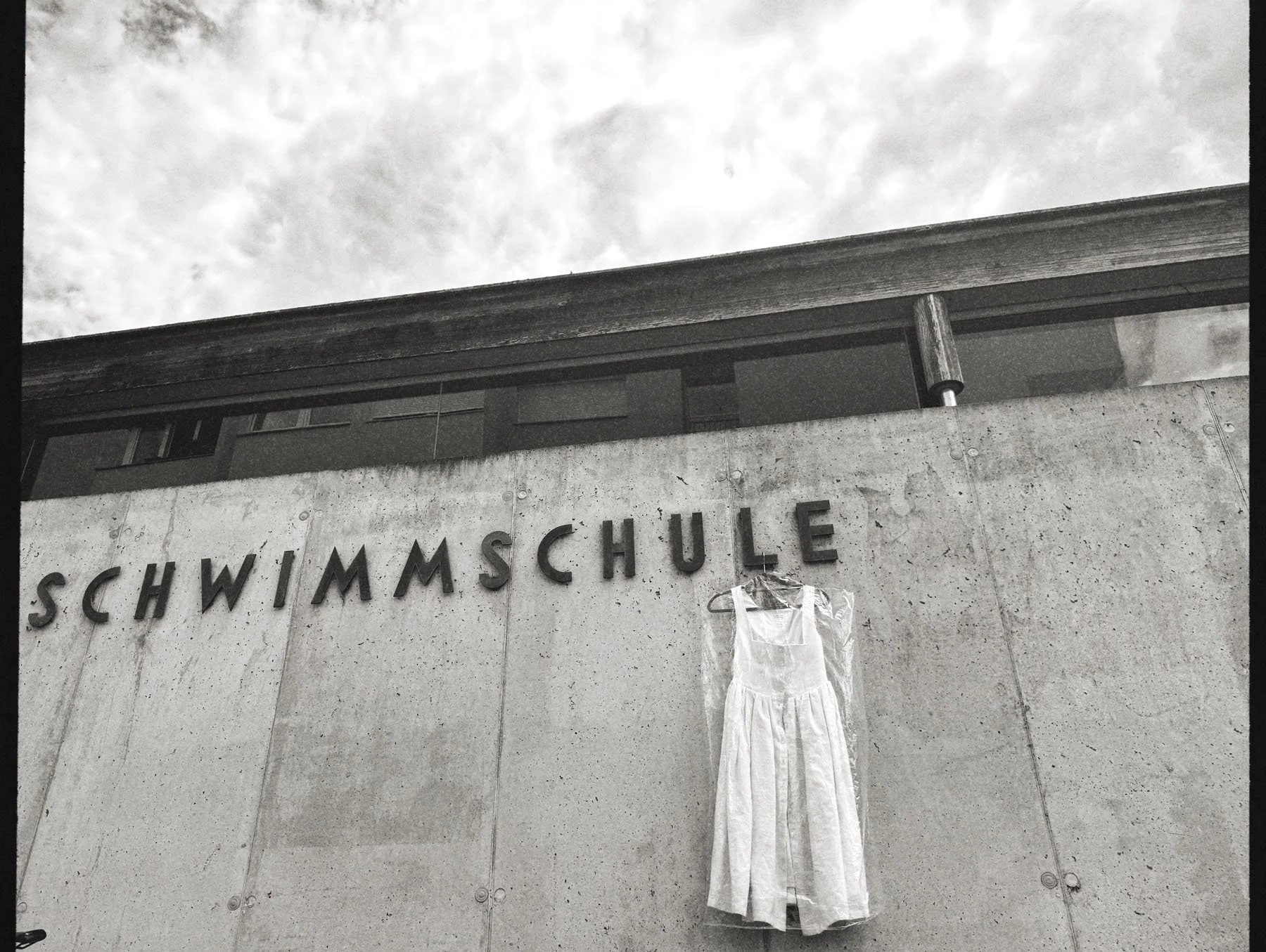 Photo of a concrete wall with the word 'Schwimmschule' on it, a dress hanging from a hook, and a building with windows and a railing in the background, taken during daytime.