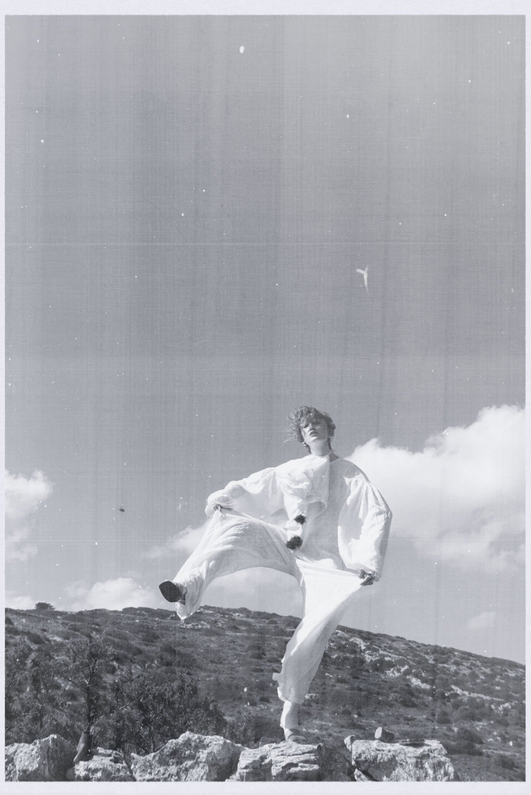 Black and white photo of a person standing on rocks outdoors, wearing a loose white dress with puffed sleeves, holding one side of the dress with one hand and lifting the leg with the other, with a sky, clouds, and birds in the background.