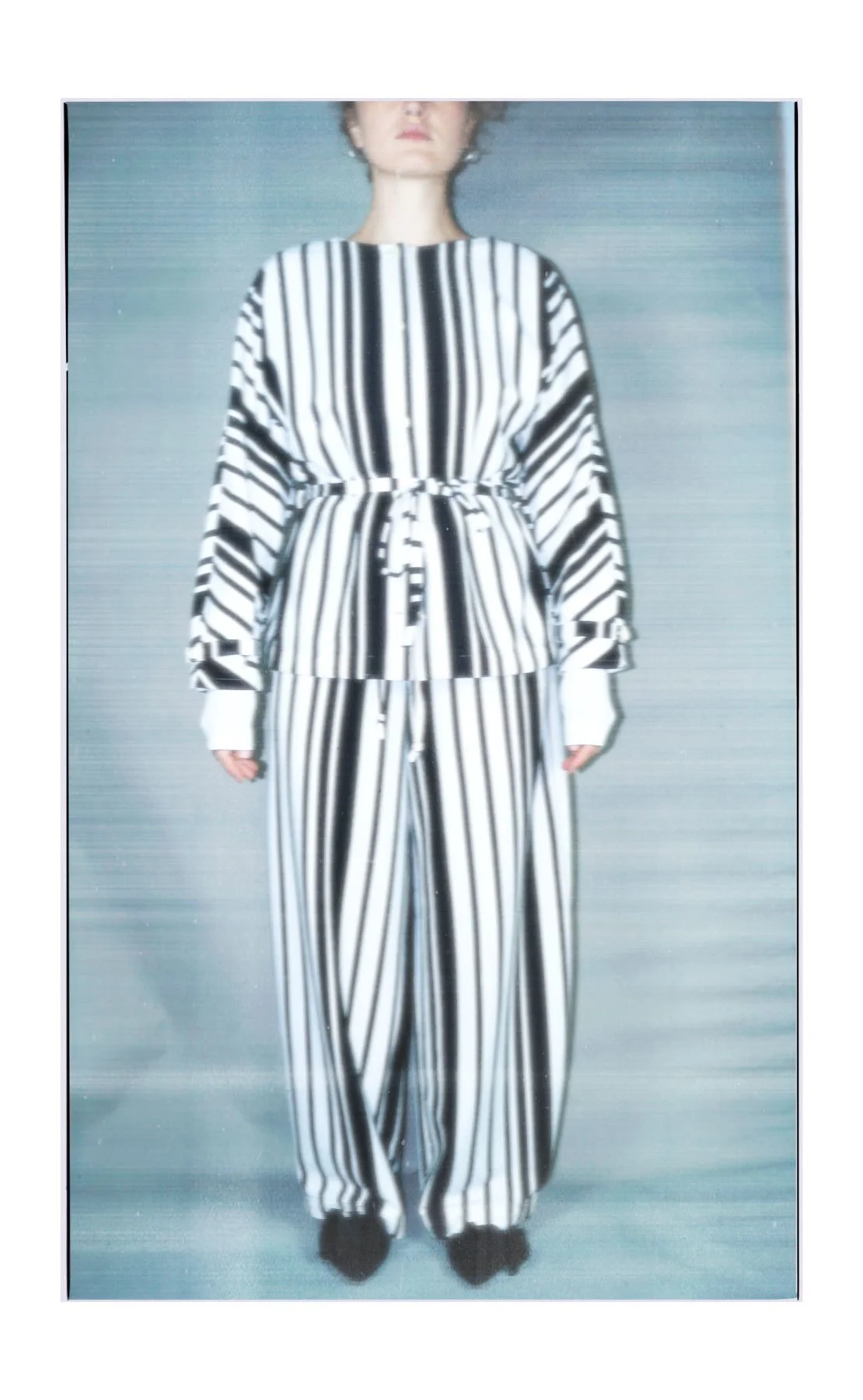 A model on a runway wearing a black and white vertically striped outfit, consisting of a top with a drawstring waist and wide-leg pants.