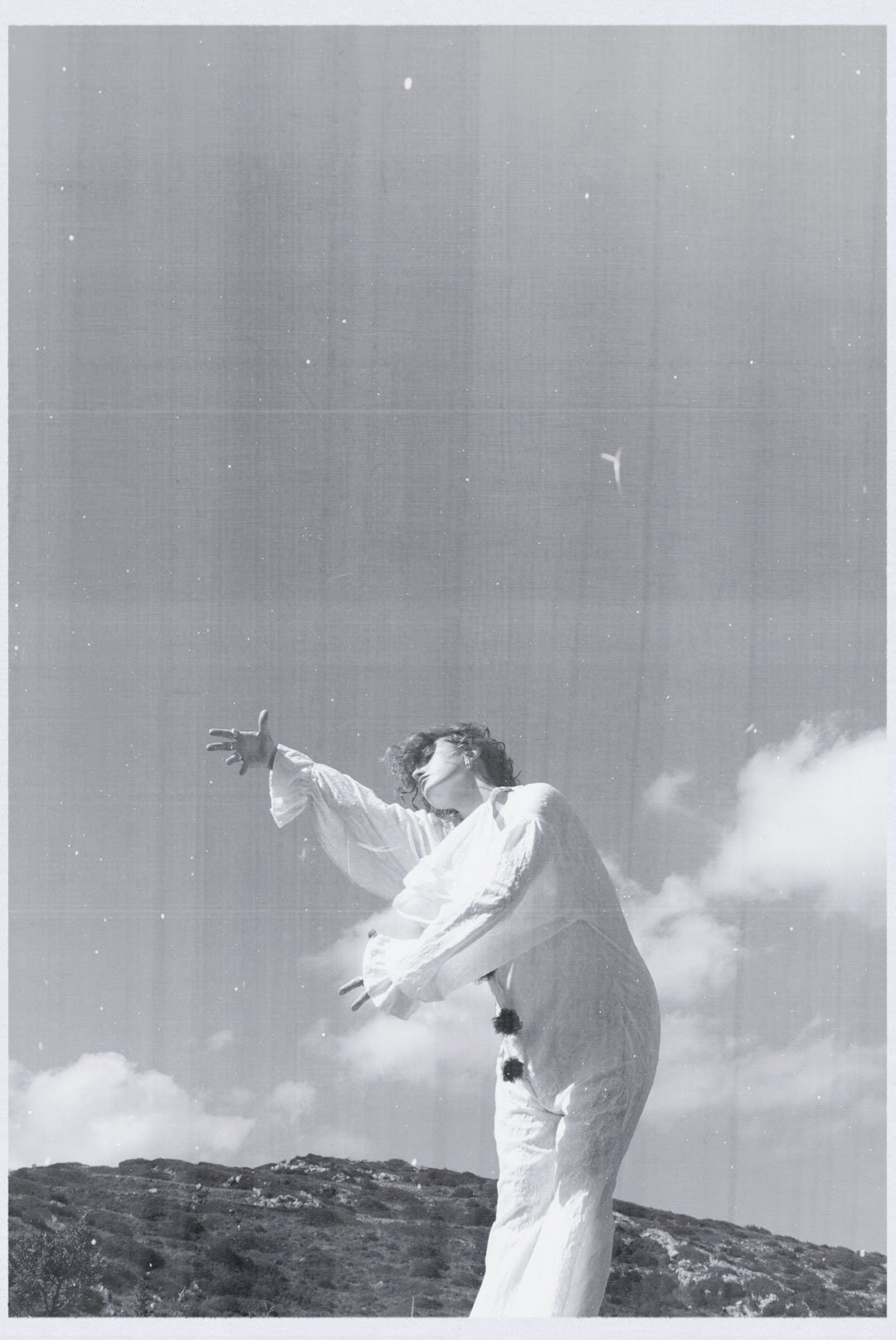 A person in a white outfit standing outdoors on a hill, with their arm extended and head tilted back, under a clear sky with scattered clouds, captured in black and white.