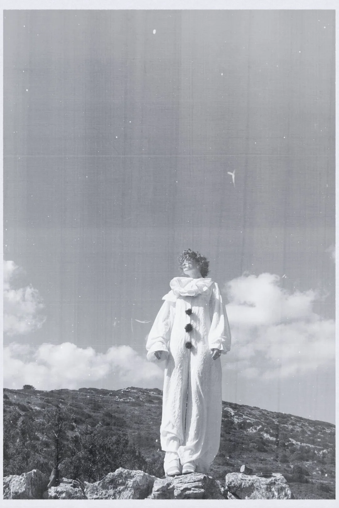 Person standing on rocky terrain outdoors, wearing a white outfit with pom-pom details, short curly hair, with clouds and a plane in the sky in the background.