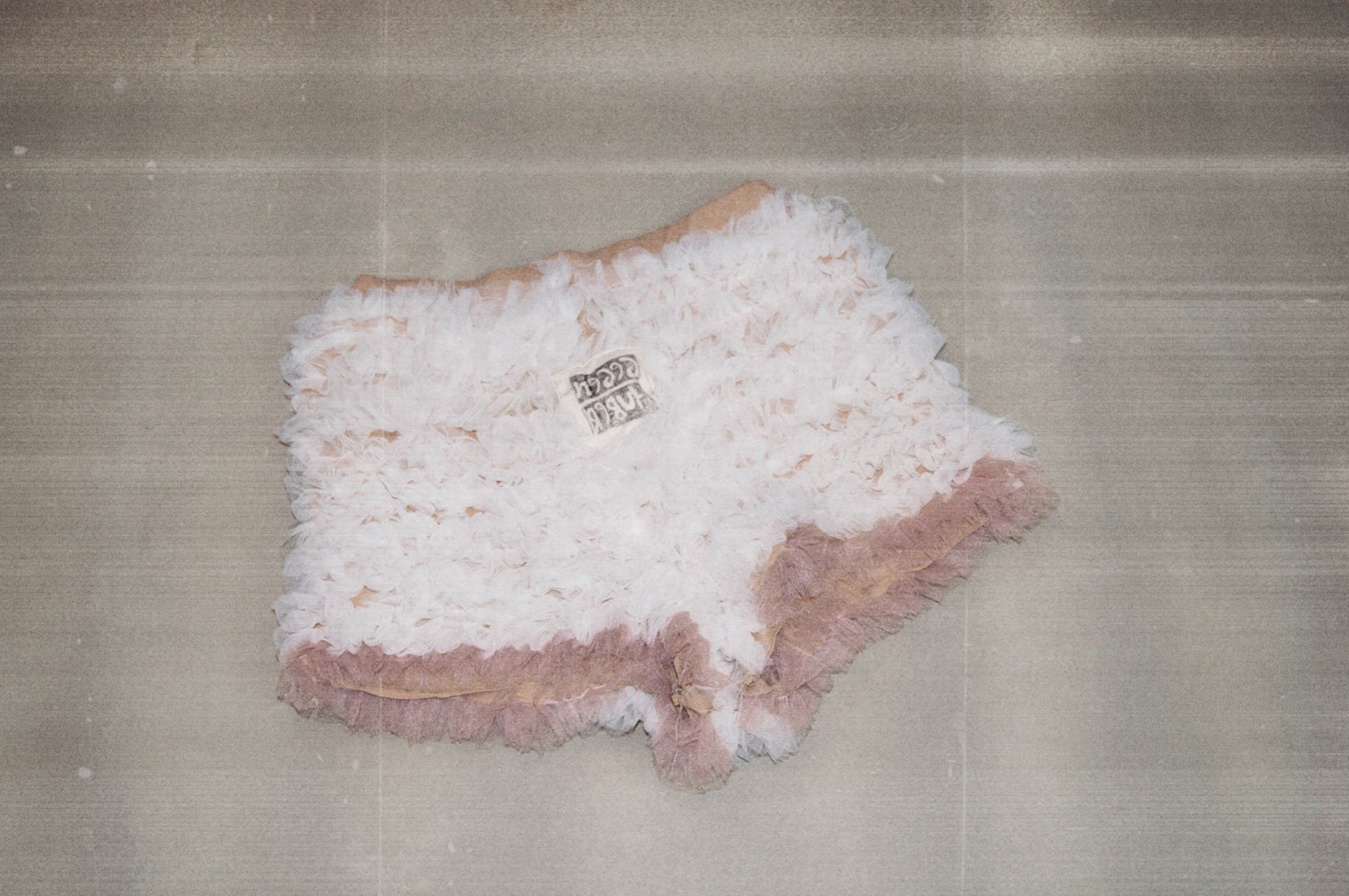 A plush toy resembling a llama or alpaca, with white fur and a beige face, lying on a gray surface. The toy has a rectangular tag attached with text.
