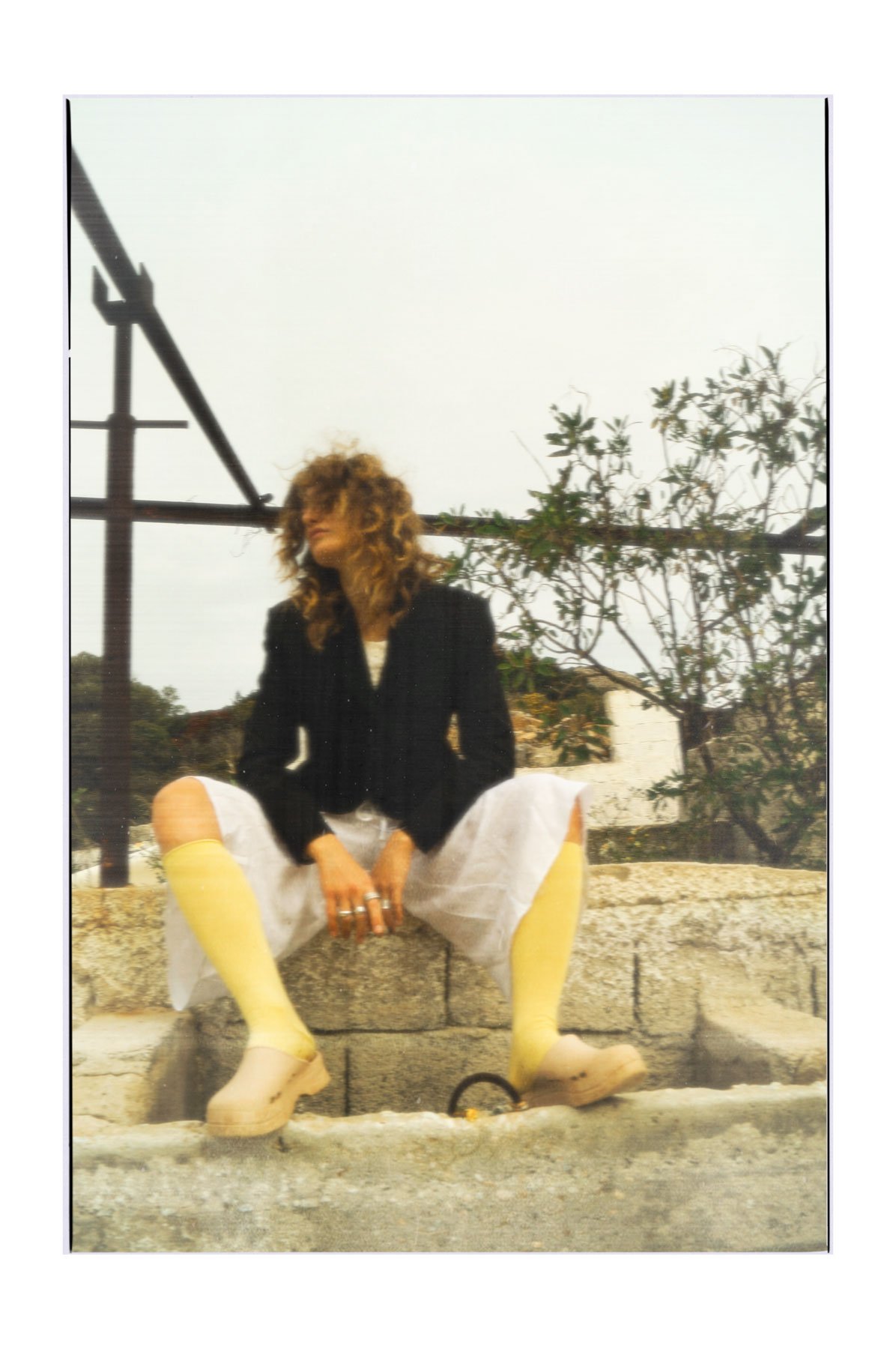A woman with curly hair dressed in a black blazer, white pants, and yellow boots sitting on a stone ledge outdoors, with trees and a cloudy sky in the background.