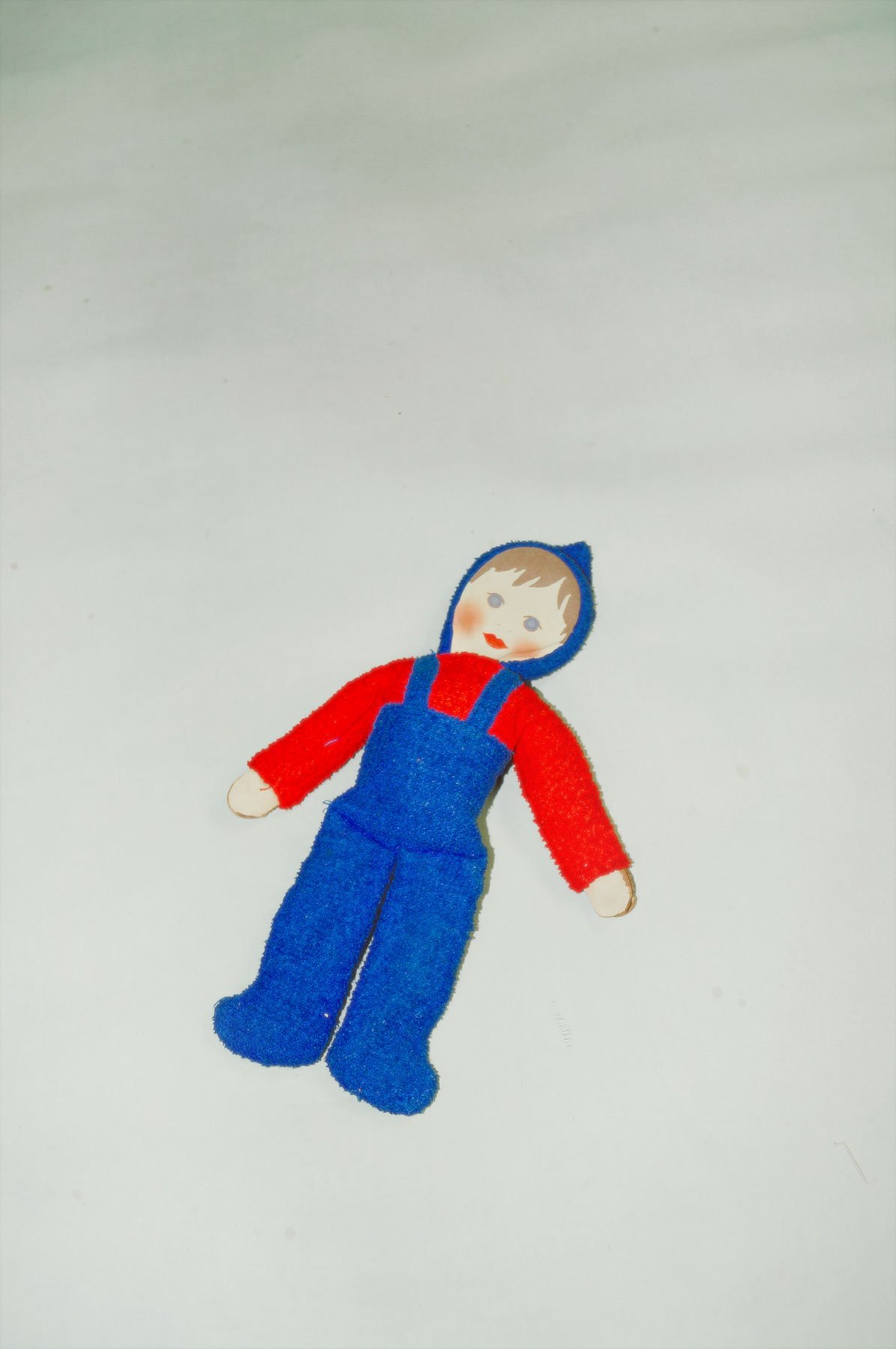 A handmade doll dressed in a red shirt, blue overalls, and a blue hood, lying on a white background.
