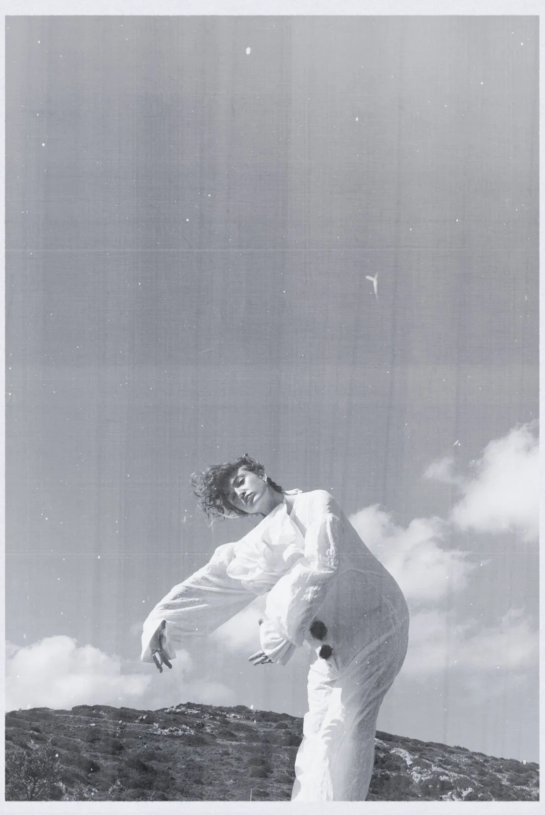 A woman with short curly hair wearing a loose white outfit standing outdoors on a hilltop with clouds and a blue sky in the background, in black and white.