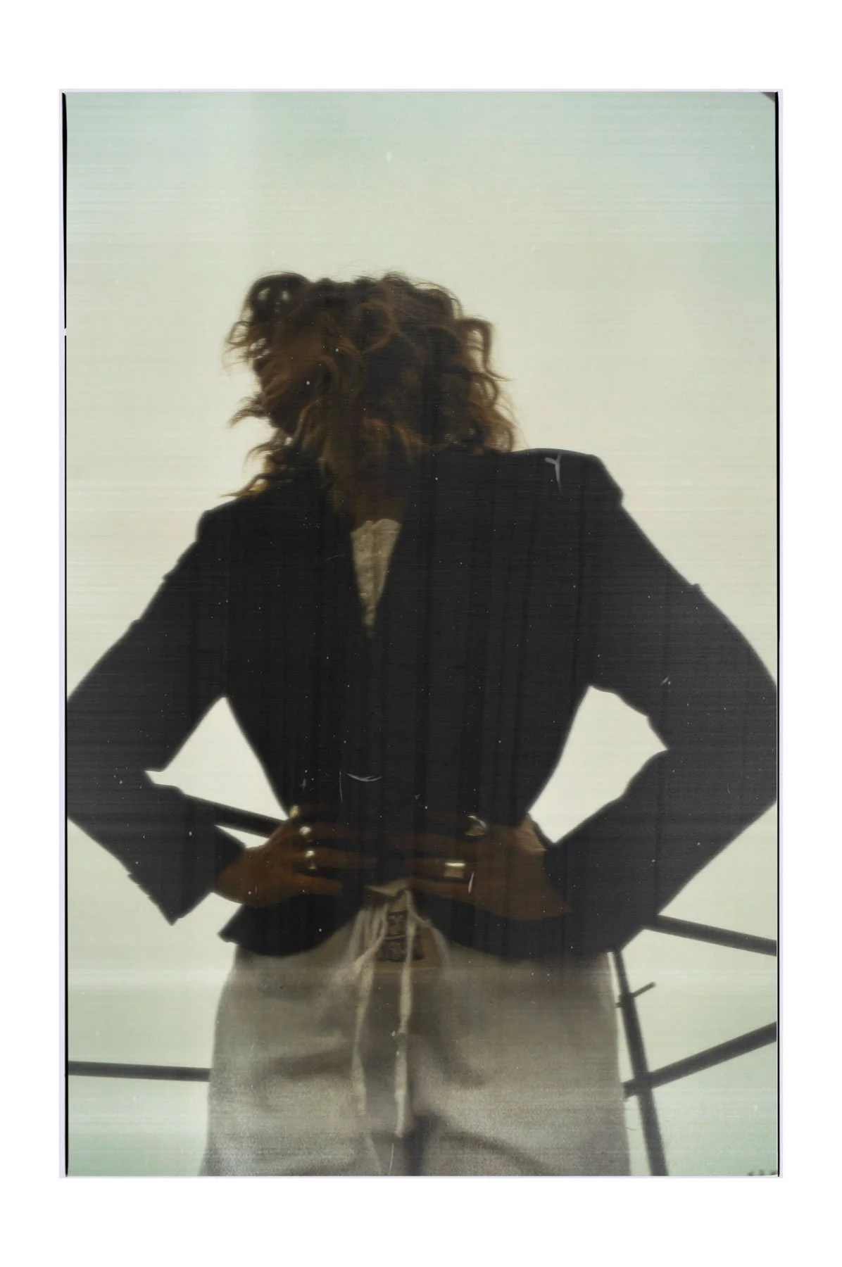 A person with curly hair wearing a black blazer and beige pants, standing with hands on hips against a plain background.