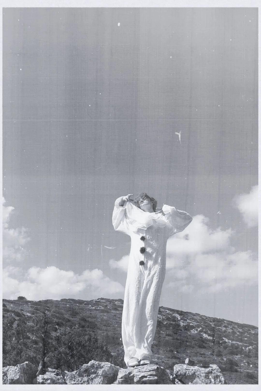 A person standing outdoors on rocky terrain under a cloudy sky, wearing a long, flowing white garment, with hands near their head and curly hair, captured in black and white.
