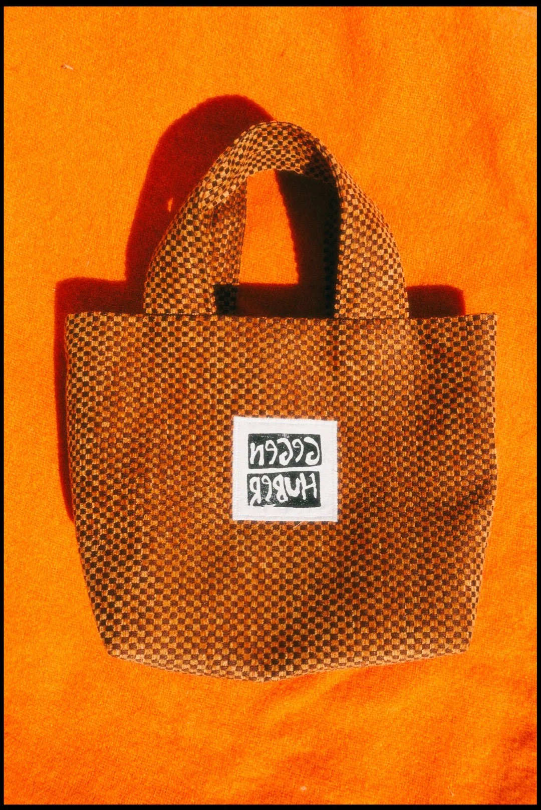 Small checkered brown and black tote bag with a white label that says 'GCCN HURR' on an orange background.