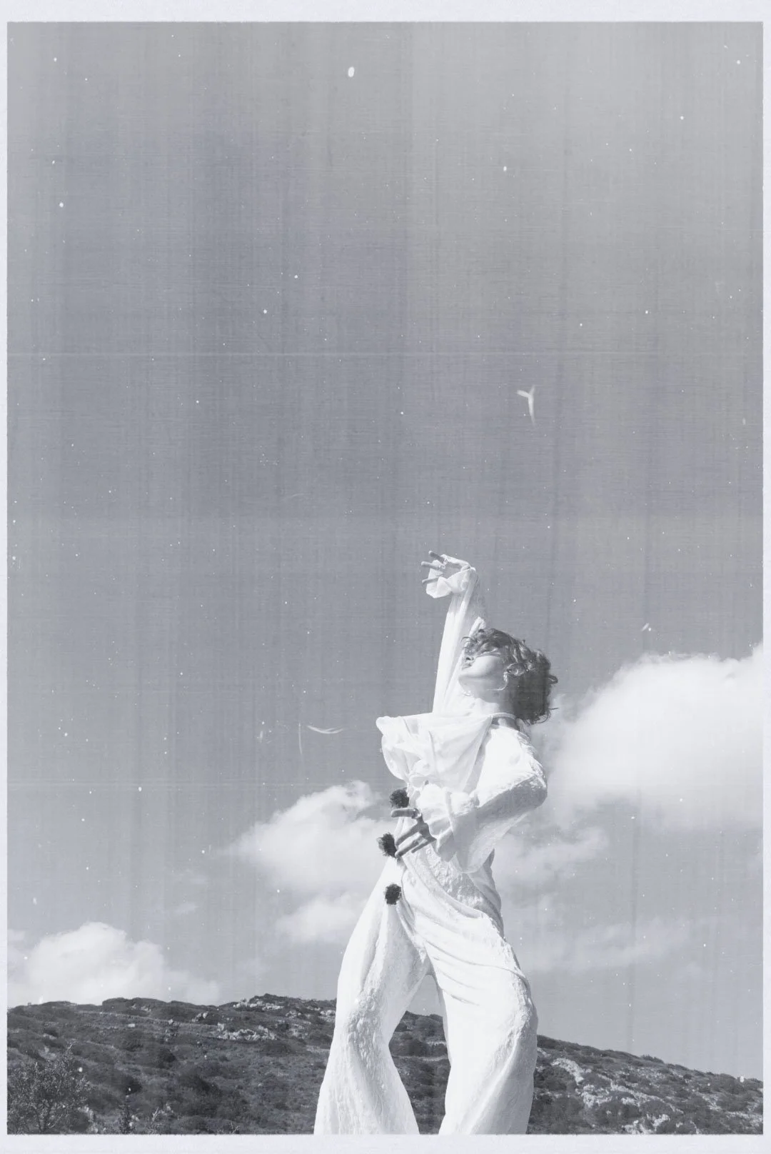 A person standing on a hilltop outdoors with arms raised and head tilted back, surrounded by clouds or fog, wearing a flowing outfit.