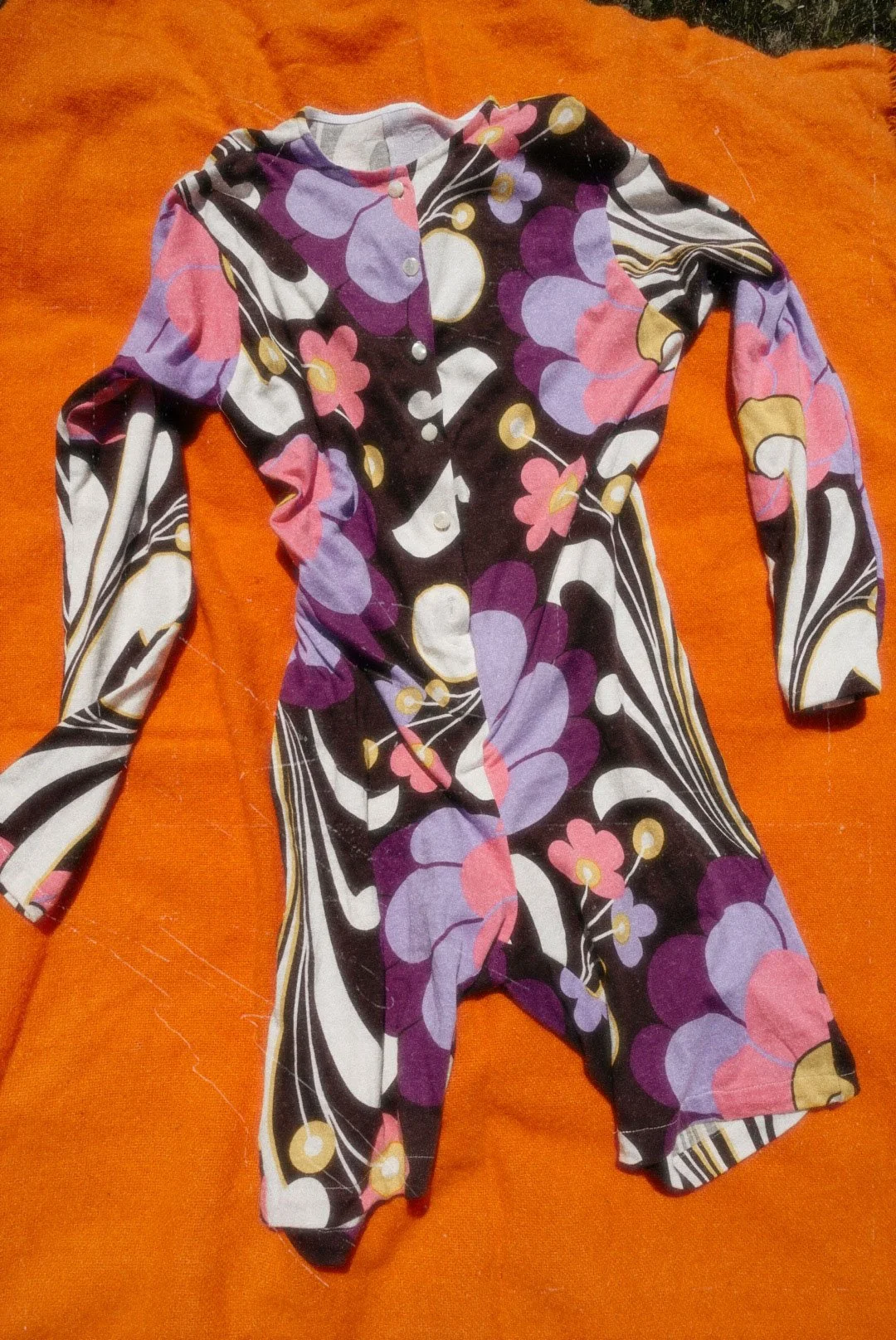A colorful, long-sleeved dress with a floral pattern in purple, pink, white, black, and yellow, laid out on an orange fabric background.