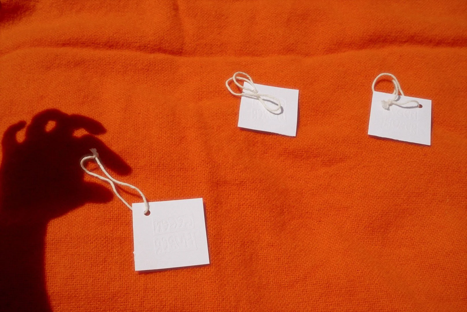 Three white tags are placed on an orange textured fabric background. The tags are attached with white strings, and the leftmost tag is being held by a person's hand, casting a shadow on the fabric.
