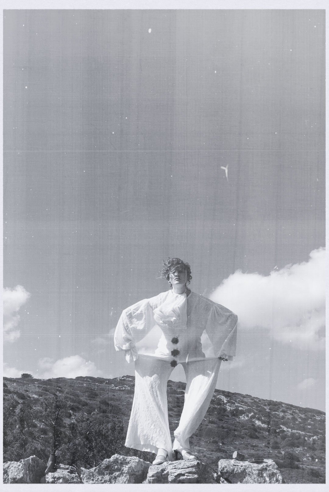 A woman standing on rocky terrain outdoors, dressed in a flowing, light-colored outfit, with her hands on her hips, facing the camera. The background features a sky with clouds and a distant hill, with a bird flying overhead.