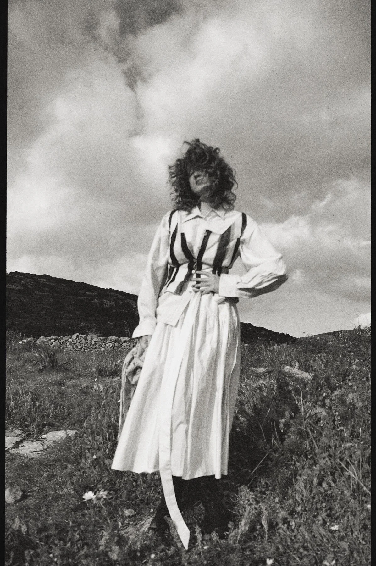 A woman with curly hair wearing a long skirt and a blouse with suspenders standing outdoors in a field with mountains and cloudy sky in the background, black and white photo.