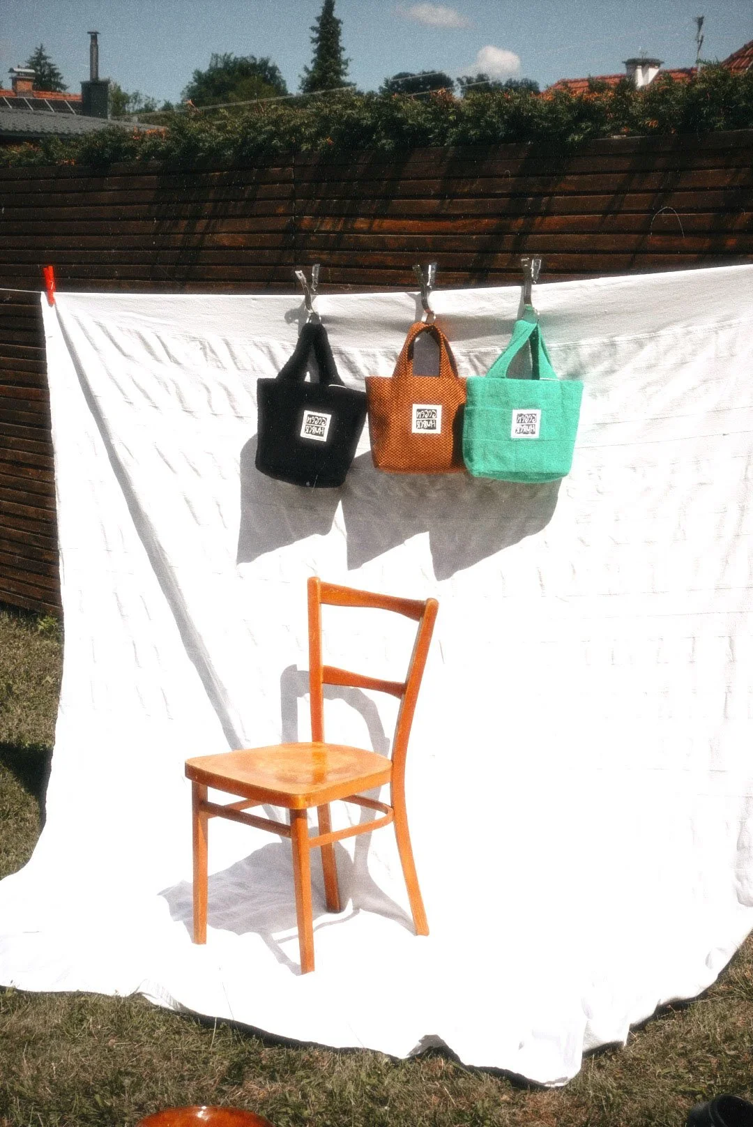 Three small bags hanging on a clothesline, black, orange, and green in color. An orange wooden chair placed in front of a white sheet backdrop with a wooden fence and rooftops visible in the background.