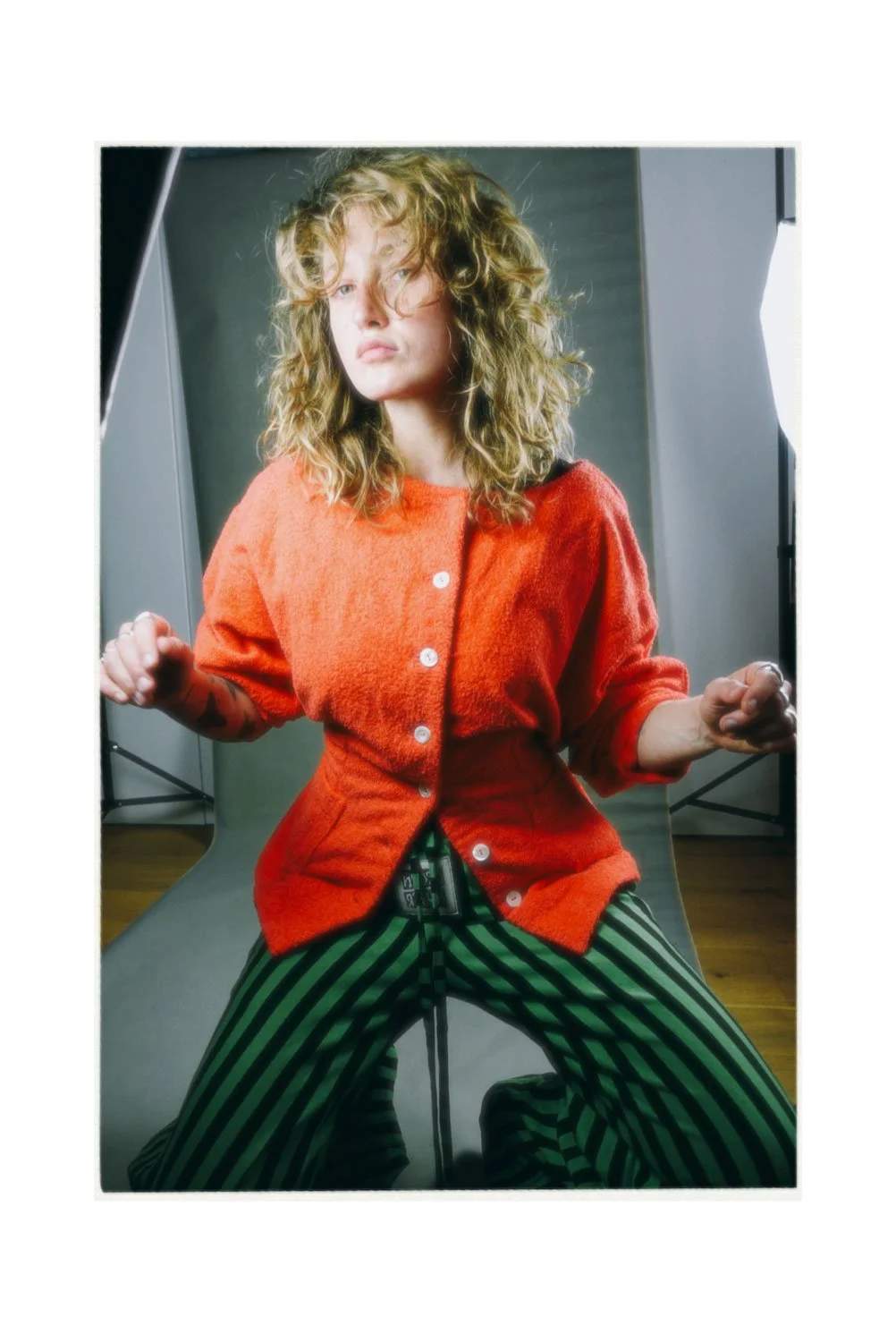 A woman with curly blonde hair sitting in front of a gray backdrop, wearing an orange cardigan and green and black striped pants, with a confident expression.