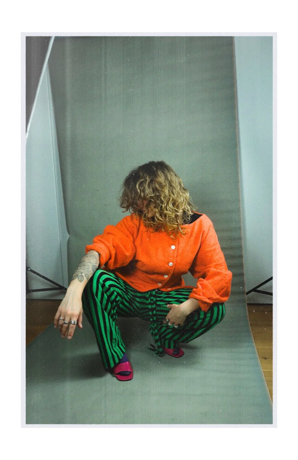 A person with curly hair squatting on a gray backdrop, wearing an orange sweater with buttons, green and black striped pants, and pink sandals.