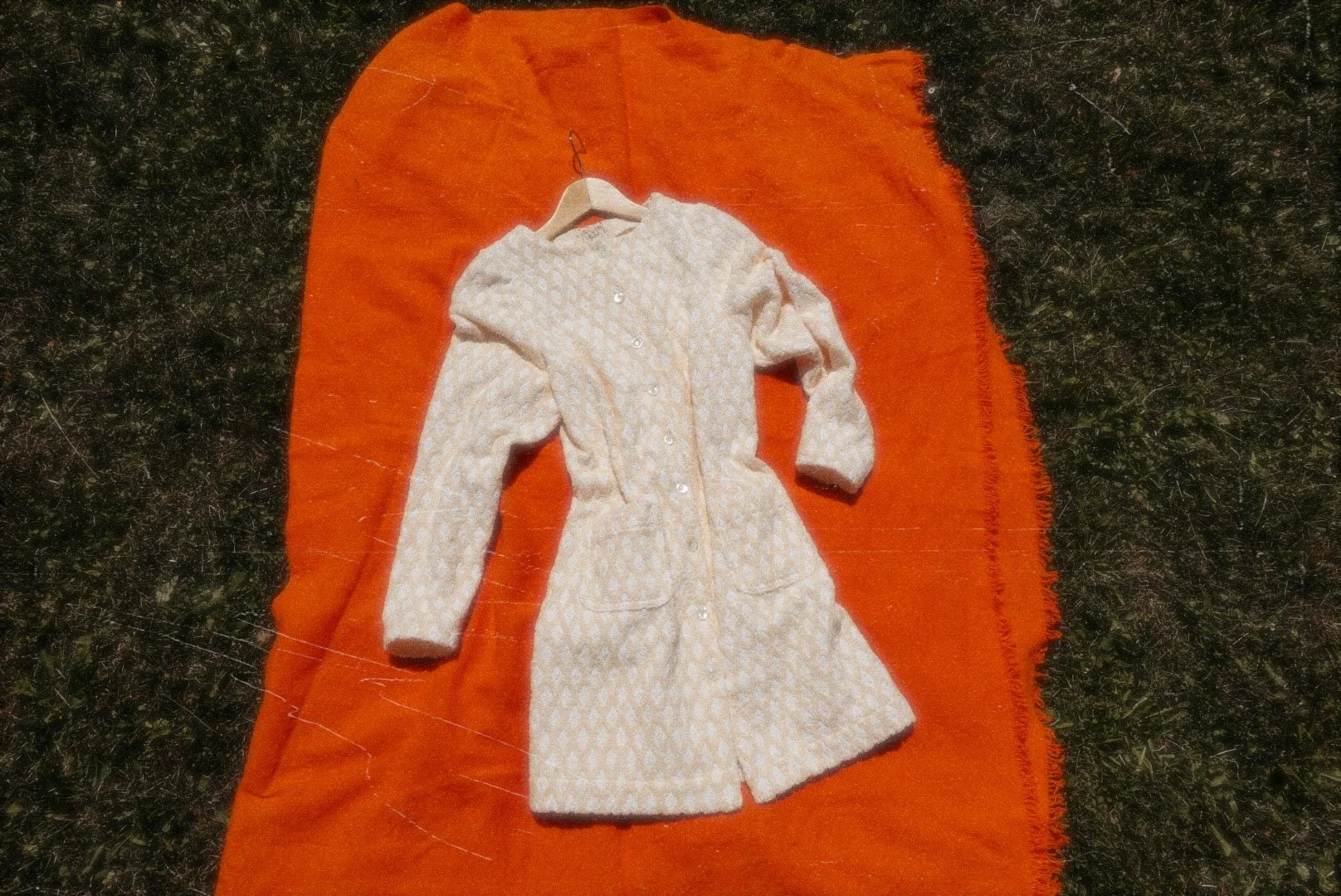 Small beige and white patterned dress with long sleeves, buttons down the front, and two pockets, lying on an orange mat outdoors.