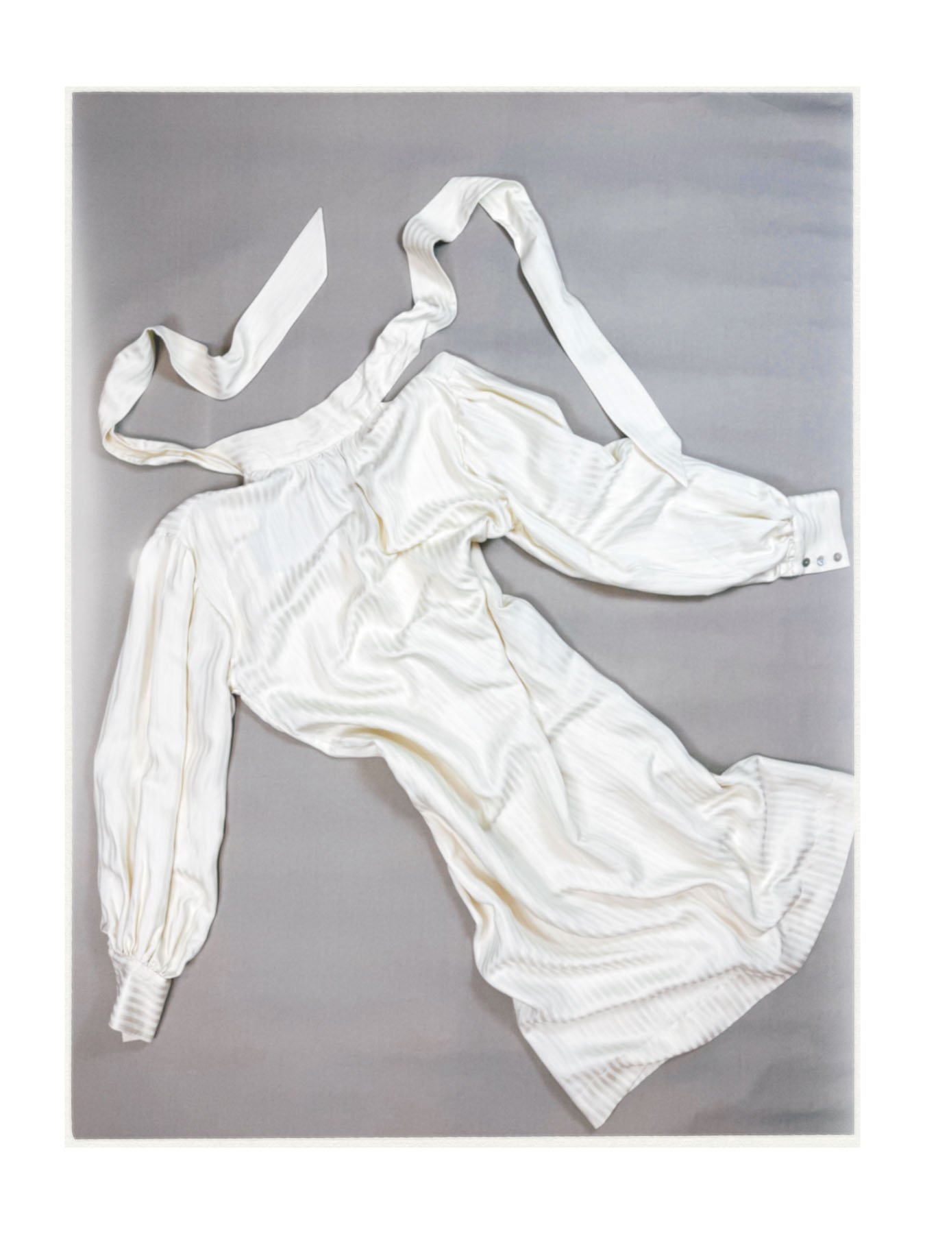 A white satin bridal wedding gown laid out flat on a gray surface, with long sleeves, a full skirt, and a long train.