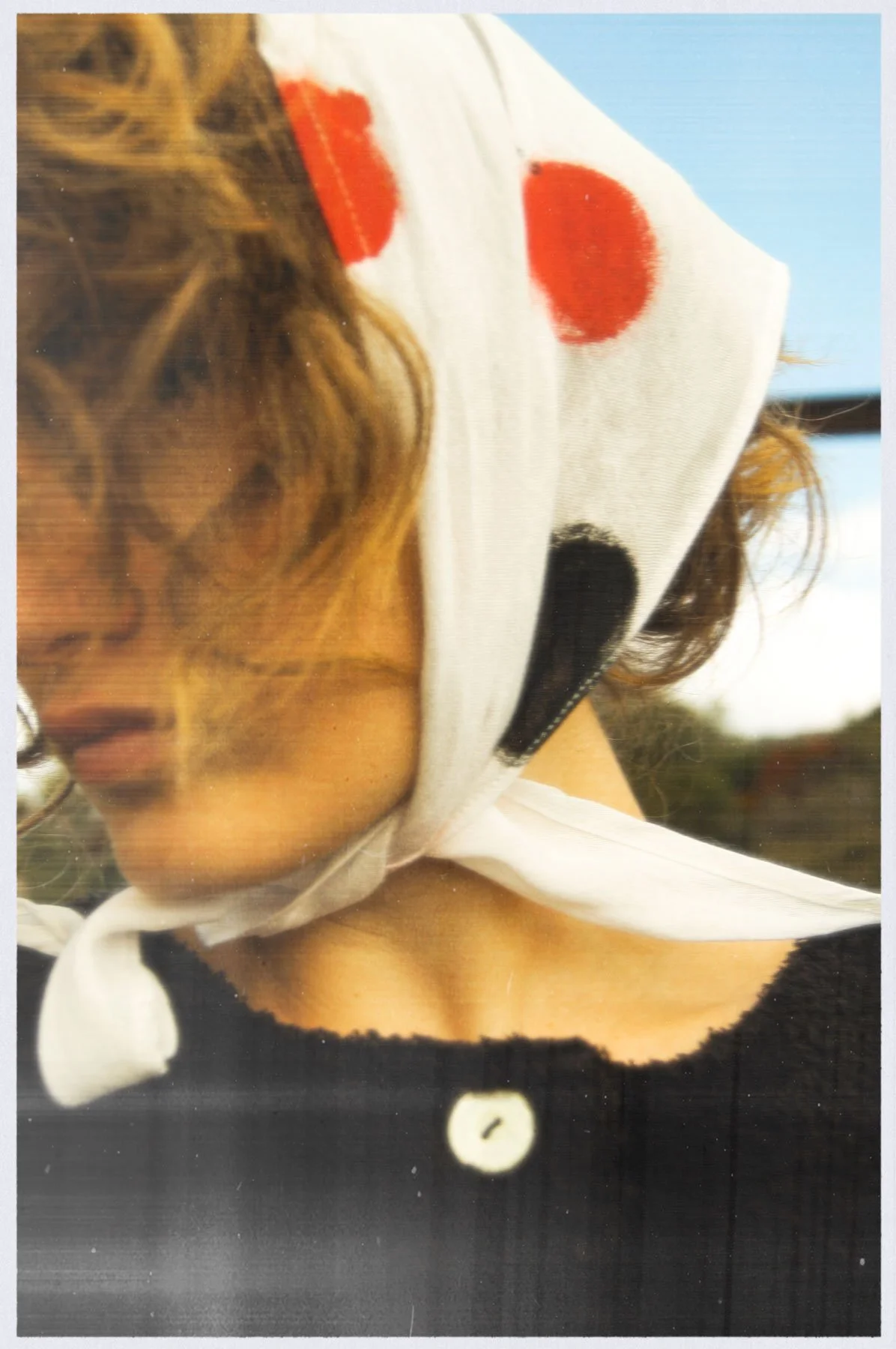 A person with curly hair wearing a white headscarf with red and black circular patterns, facing downward, outdoors with a blue sky in the background.