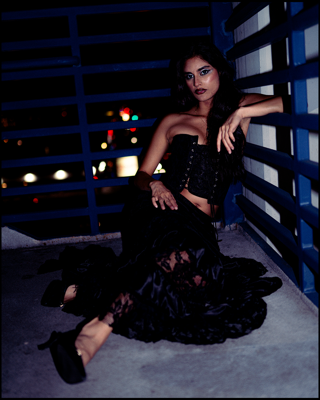 A woman in dark clothing sitting on a rooftop at night, leaning against a blue railing with city lights in the background.