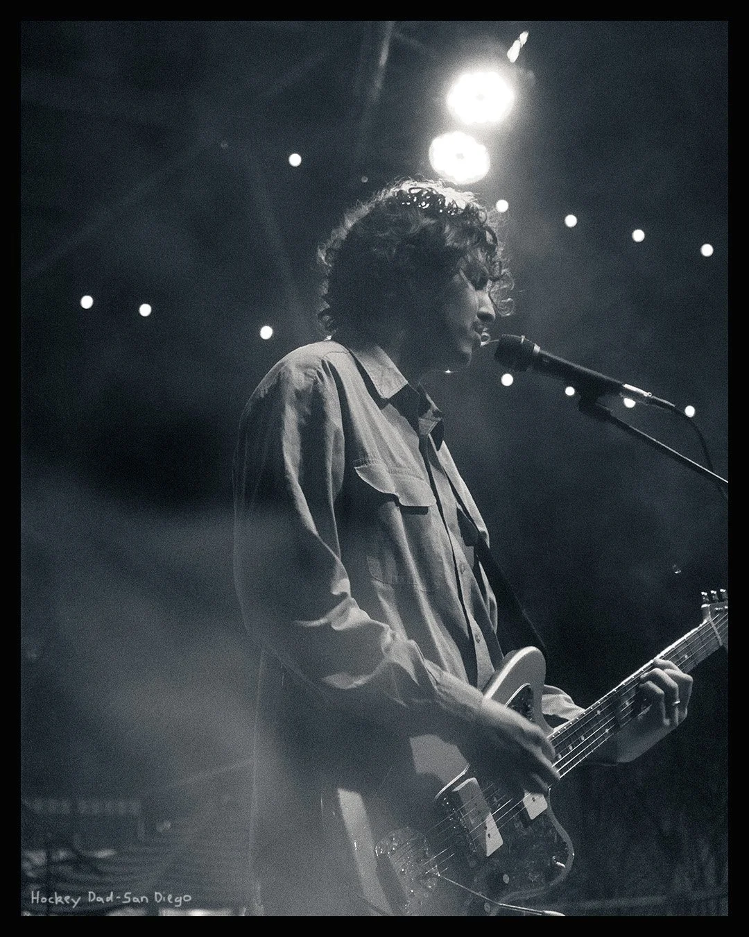 A musician with curly hair performs on stage, playing an electric guitar and singing into a microphone, illuminated by stage lights.