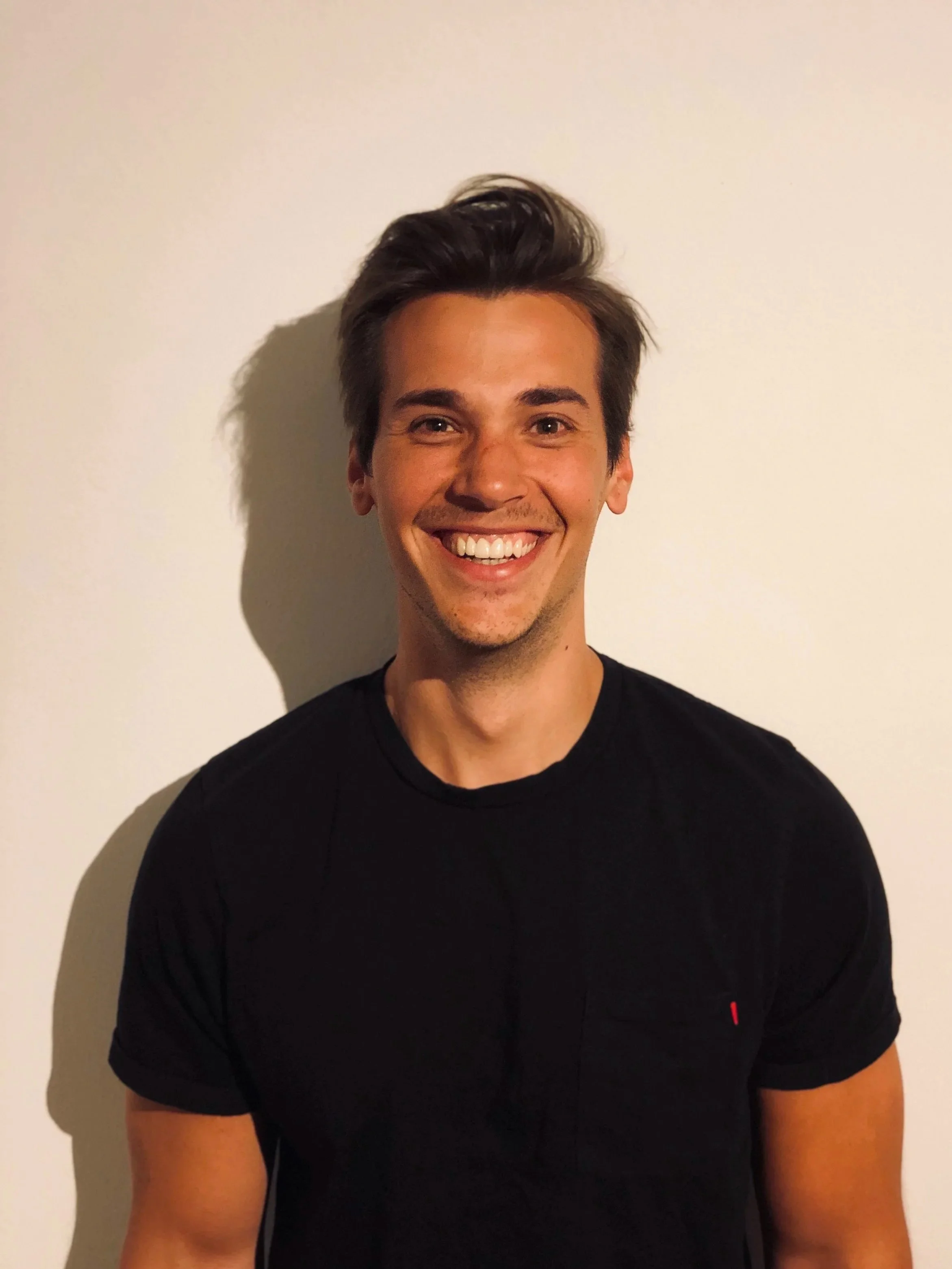 Ein junger Mann mit dunklen Haaren, breitem Lächeln und hellem Teint trägt ein schwarzes T-Shirt und steht vor einer weißen Wand.