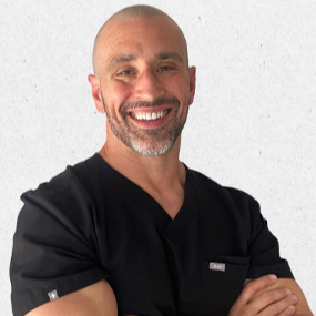 Dr. Aaron Sakevich is smiling in black scrubs against a light wall.