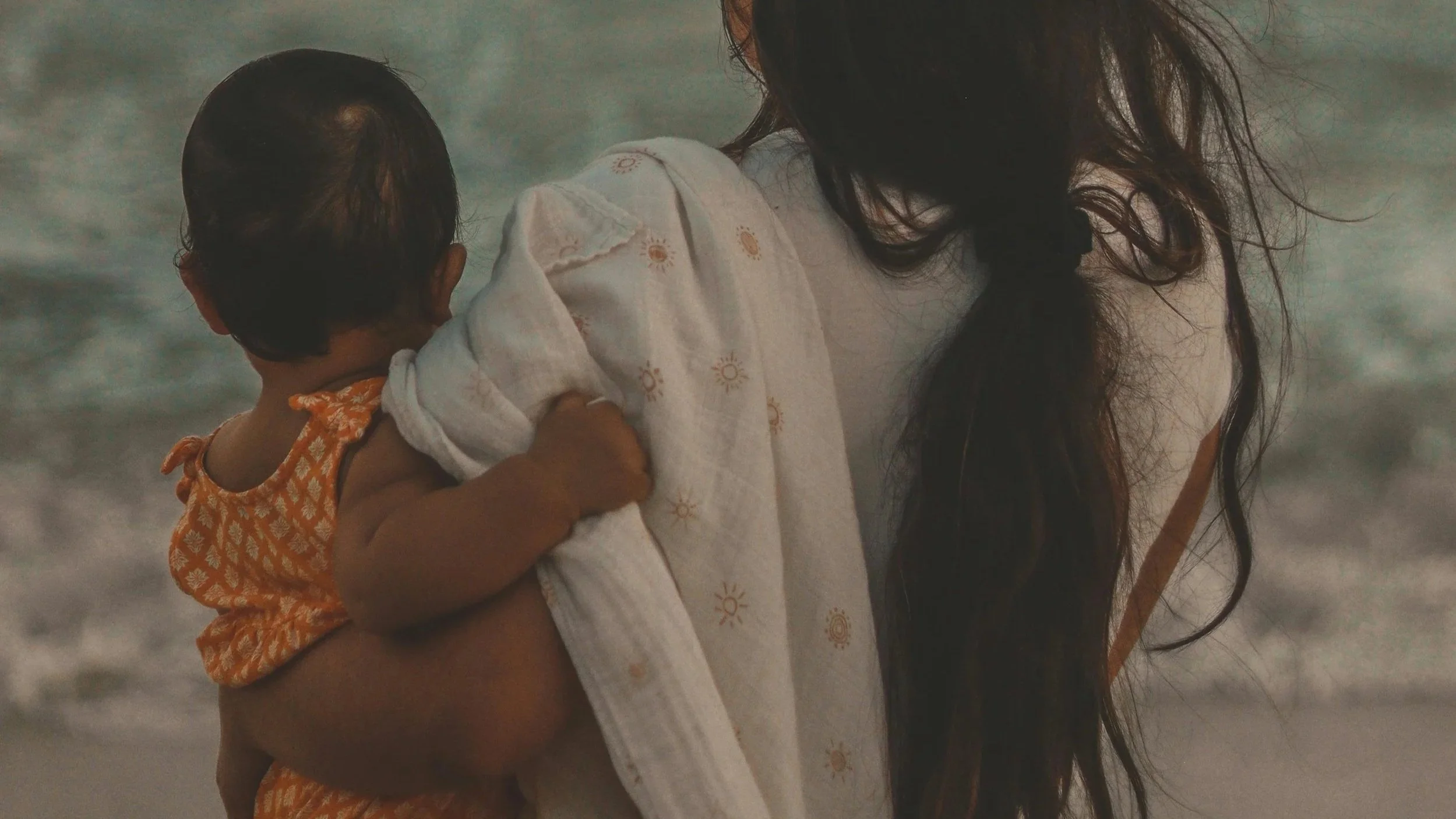 close up of the back of a mom holding baby daughter in front of the ocean