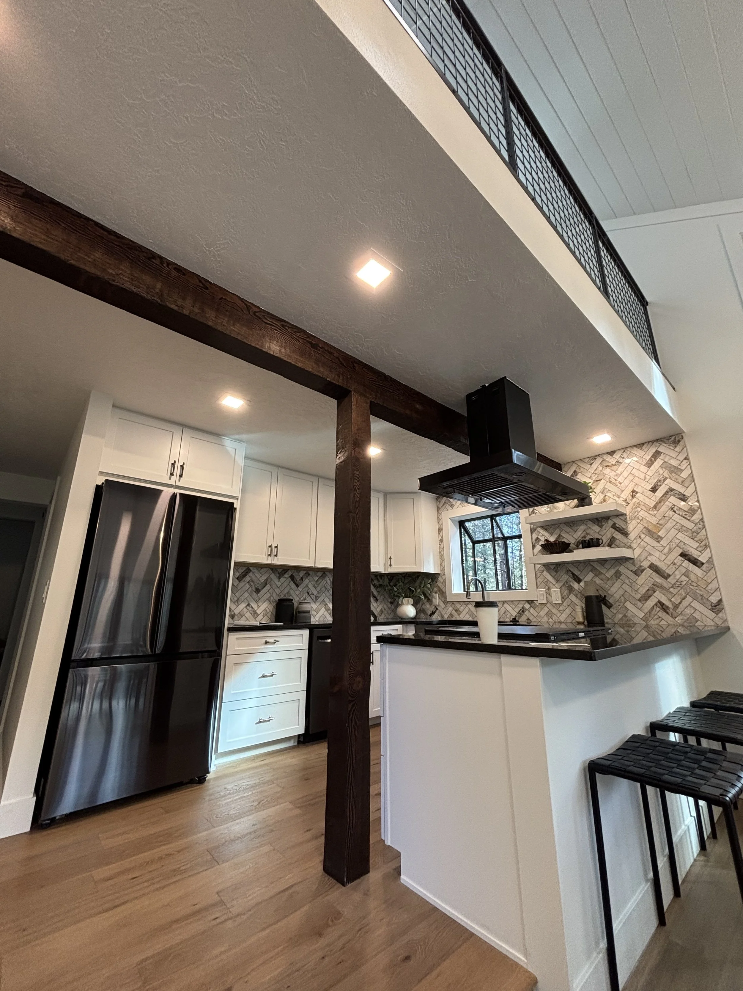 Modern kitchen with white cabinets, black appliances, a herringbone-patterned backsplash, and a wooden support beam.