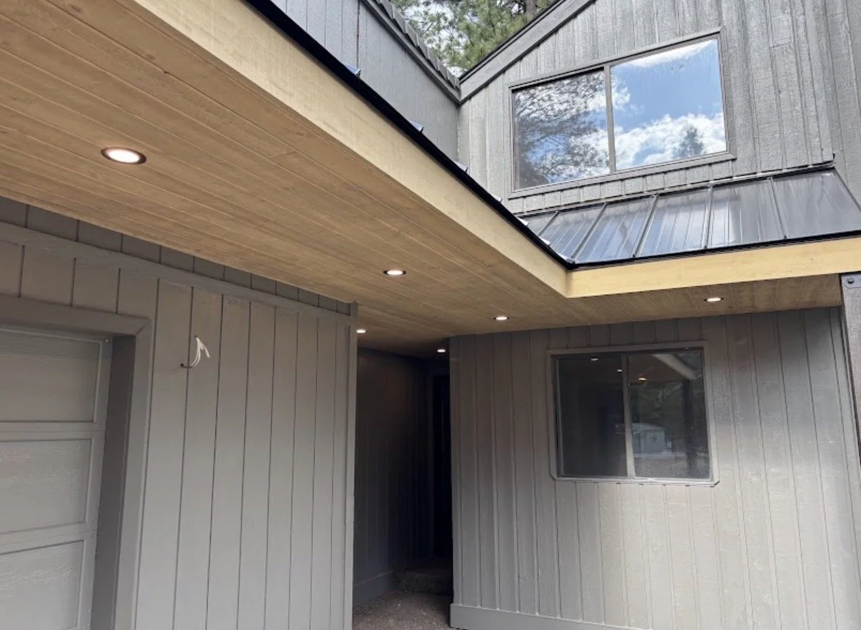 Exterior view of a modern house with gray wood siding, a garage door, and a covered patio area with recessed lighting. There is a large window on the upper level reflecting the sky and trees.