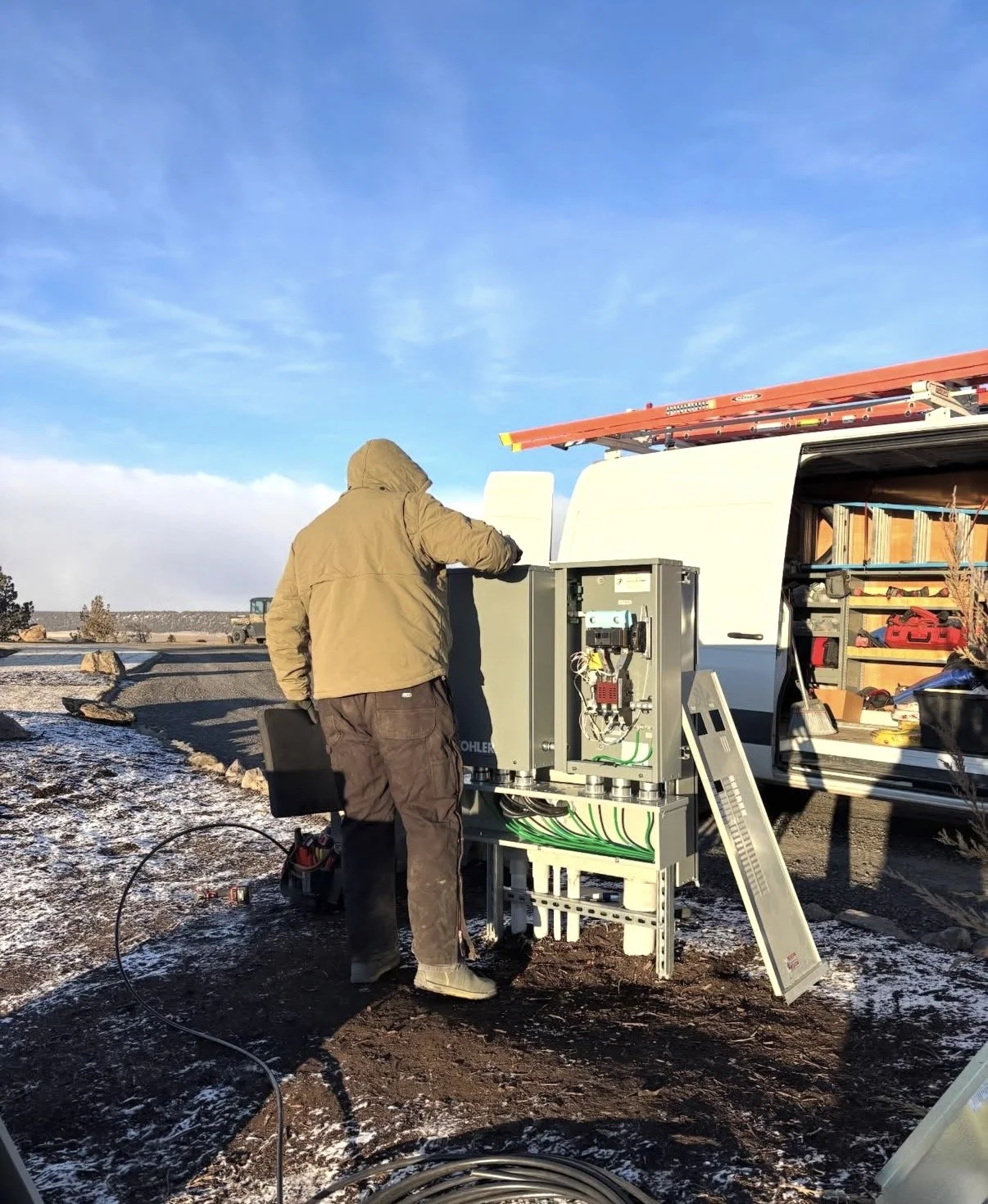 A person in a brown jacket and pants working on electrical equipment outside near a white van with an open side door. The van contains tools and supplies, with a ladder on top. 