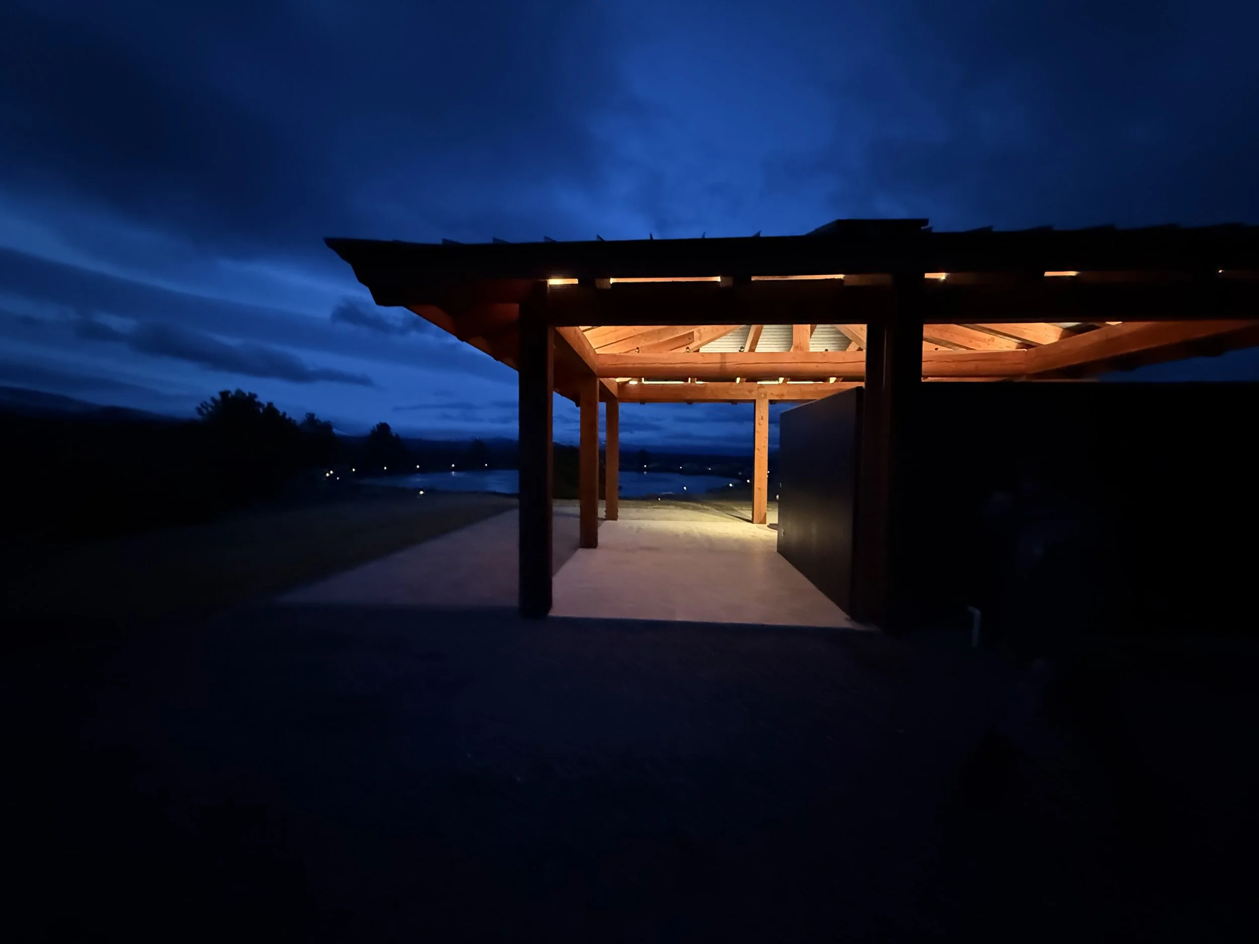 A wooden shelter illuminated at dusk, with a roof supported by wooden beams, overlooking a landscape with water and distant trees under a dark, cloudy sky.