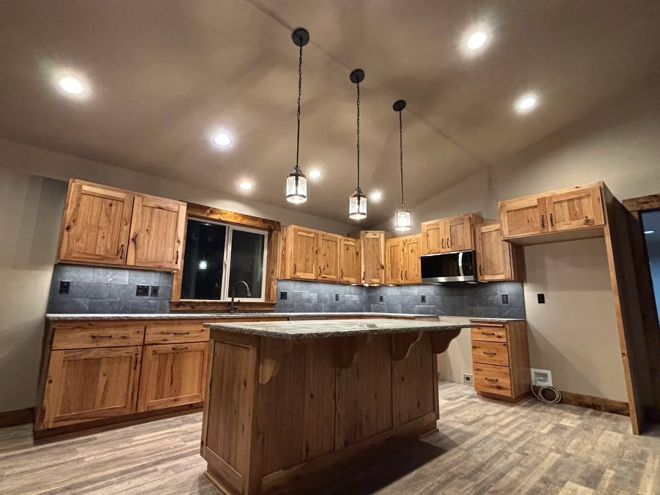 Kitchen with wooden cabinets, granite island, black tiled backsplash, and pendant lighting.