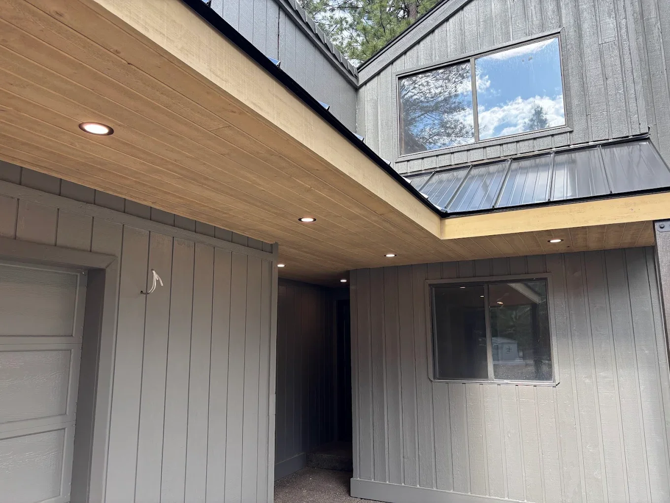The image shows the exterior of a modern house with dark gray vertical siding, a wooden ceiling with built-in lights in the covered porch area, and a large window reflecting the sky and trees. There is also a smaller window and part of a garage door 