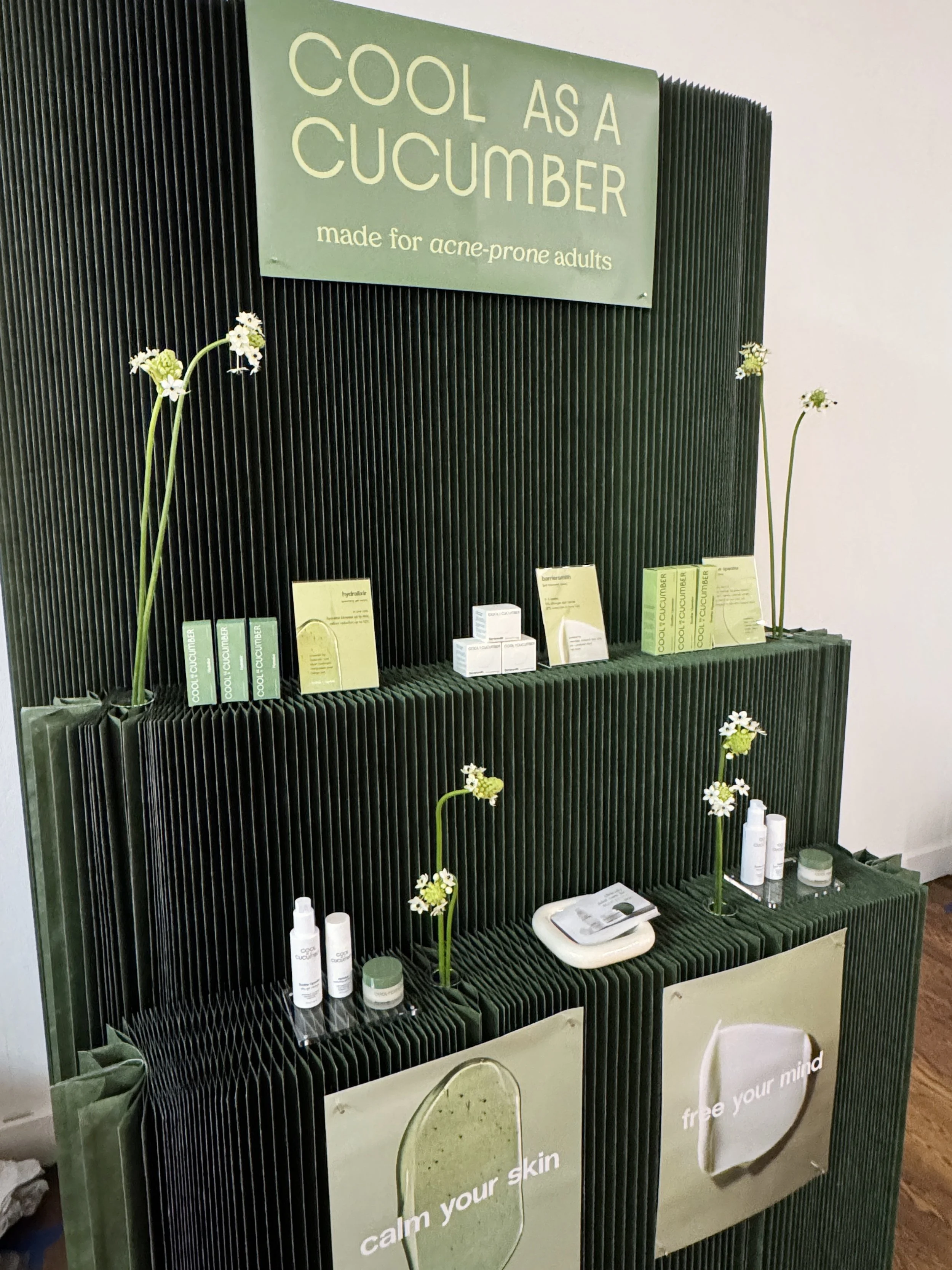 Display stand with skincare products and a sign that reads 'Cool as a Cucumber, made for acne-prone adults,' decorated with white daisies.