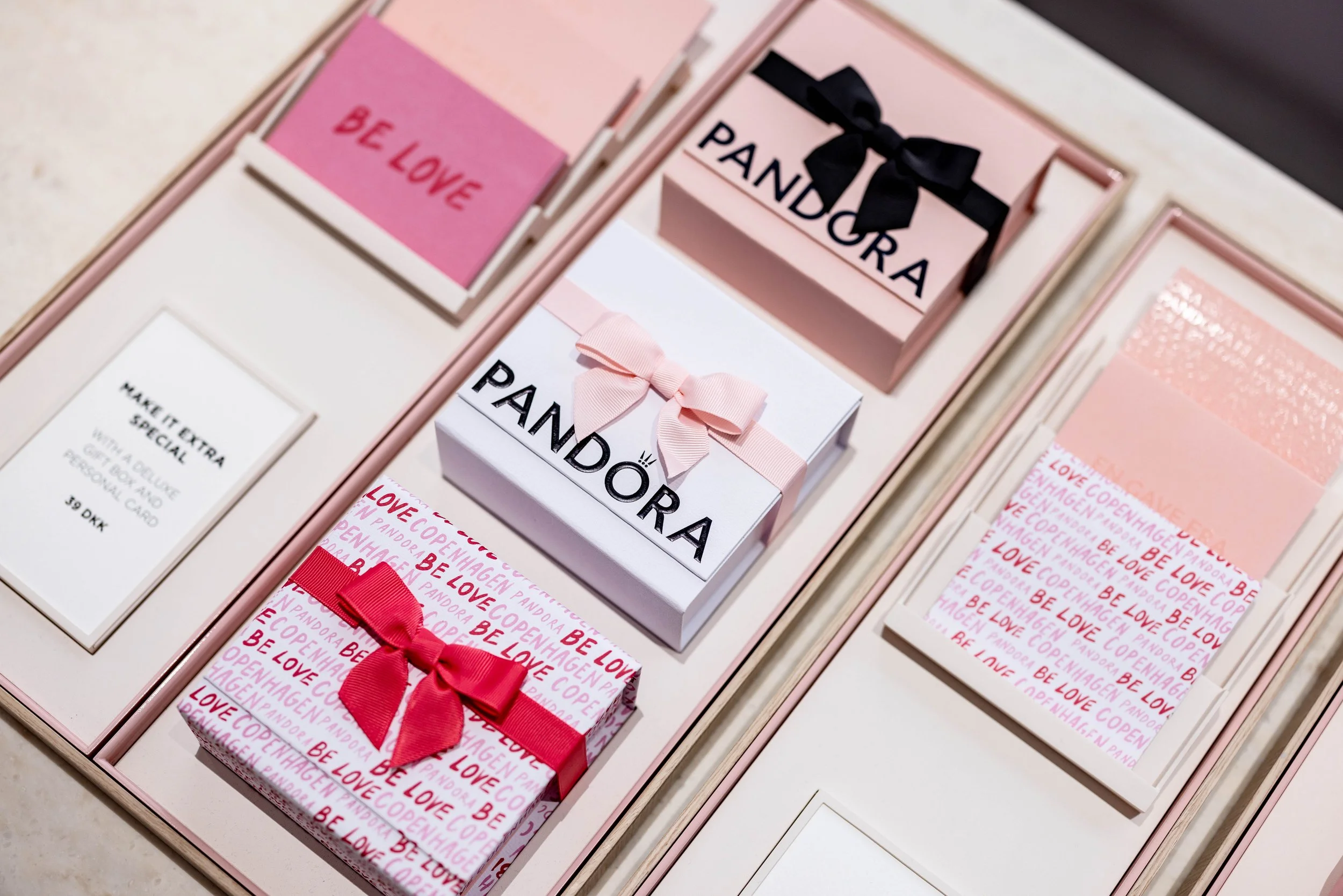 A pink gift box with a red ribbon and 'Be Love' written on a pink card inside a gift box, alongside a white gift box with a pink bow and 'Pandora' branding, and a pink patterned card with 'Be Love' text repeated.